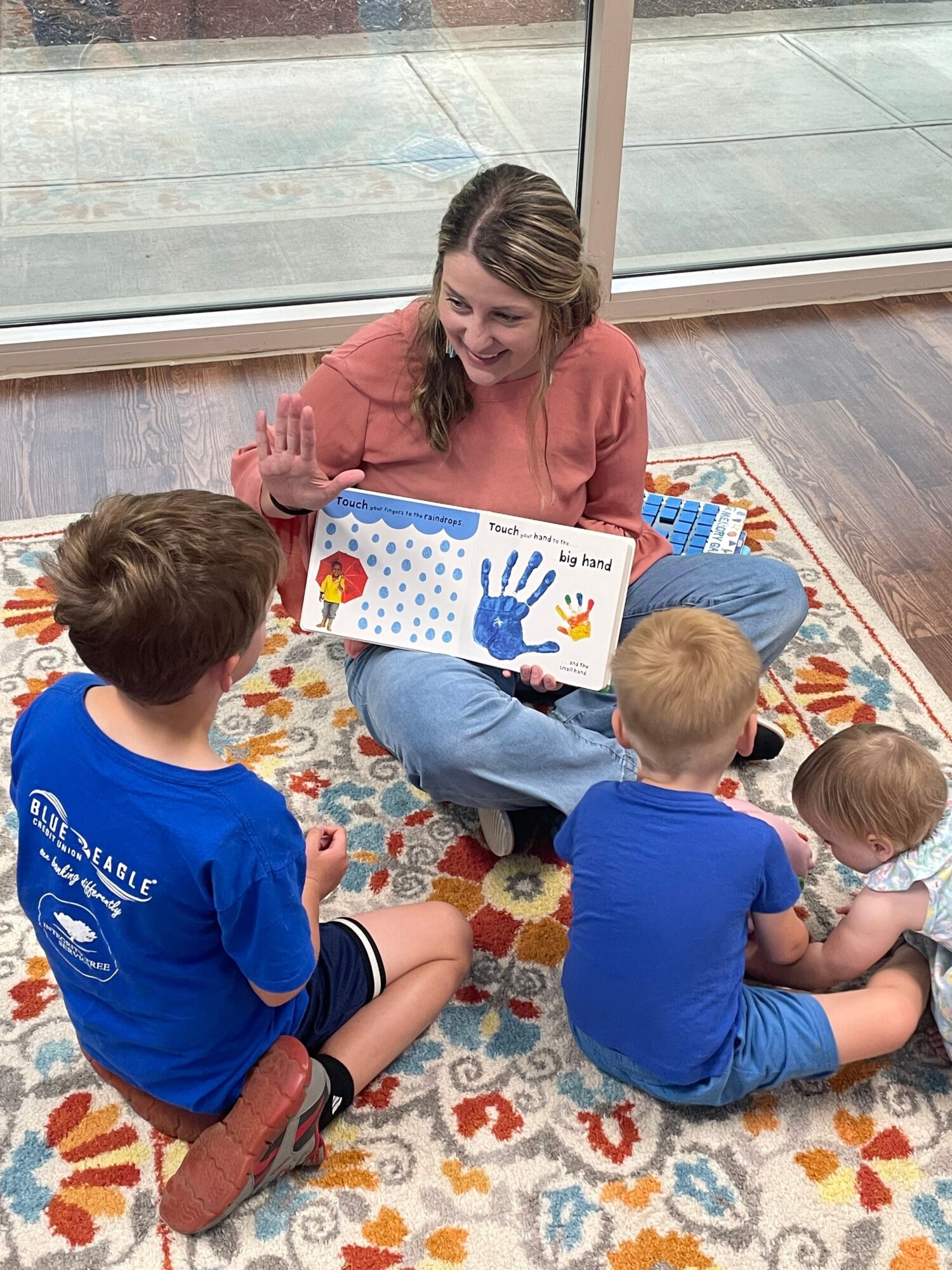 Woman reading a colorful book to two children sitting on a patterned rug near a window.