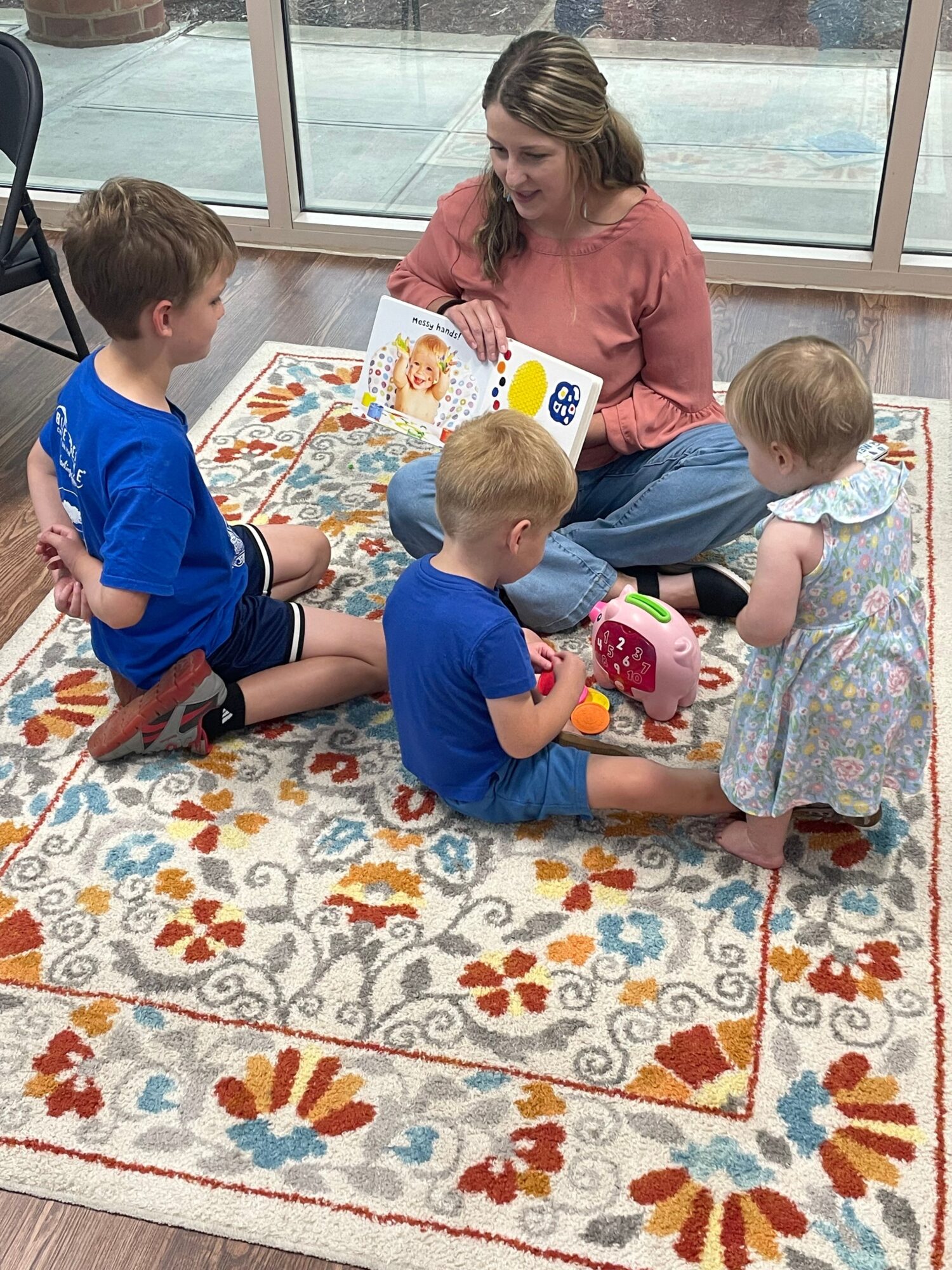 Adult woman with three children sitting on a colorful patterned rug near a window, engaging with toys and books.