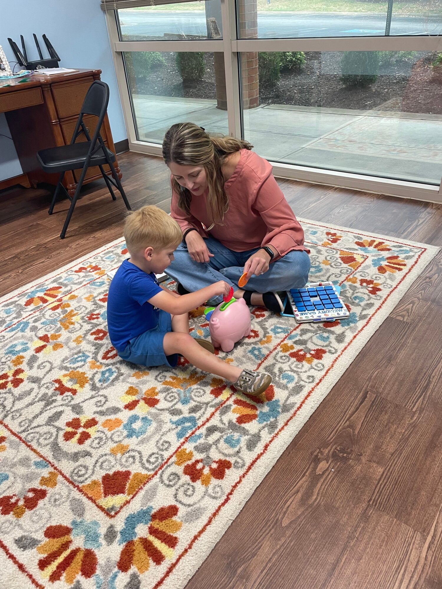 Two children sitting on a colorful patterned rug near a glass door, playing with toys indoors.