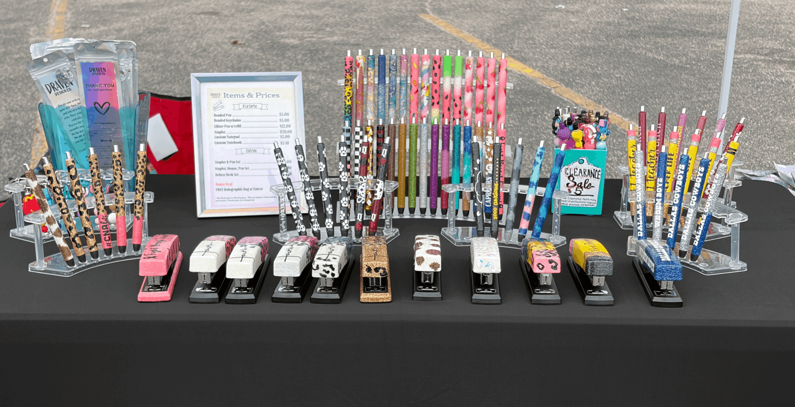 Display of colorful pens, markers, and stationery items on a table with a black cloth, including a framed sign and small containers.