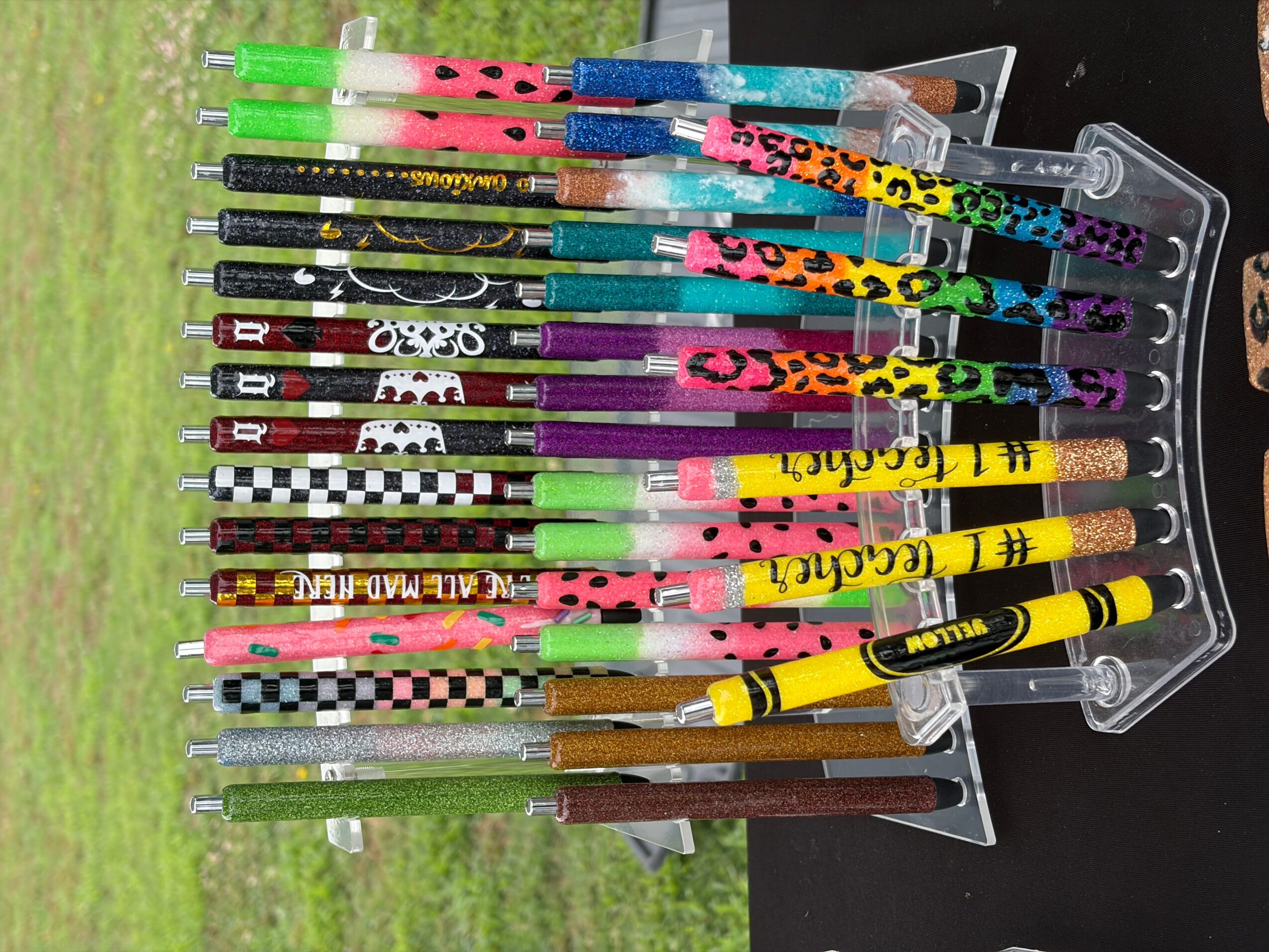 Colorful pens and markers in a plastic holder on a black surface, with a grassy background visible outside.