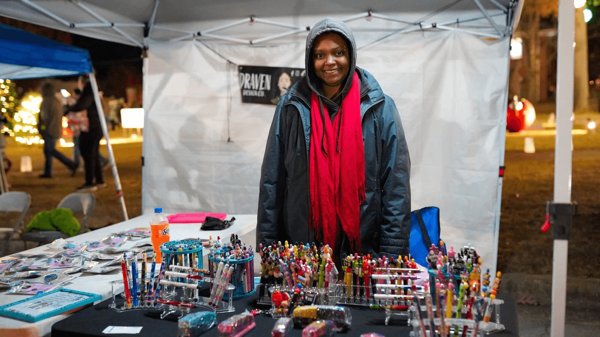 Person standing behind a table with colorful pens and stationery at an outdoor market stall, smiling, wearing a red scarf and dark jacket.