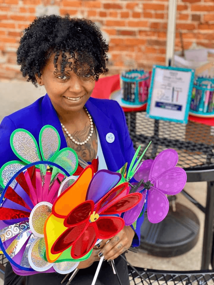 Woman holding colorful paper flowers, wearing a blue blazer and pearl necklace, standing outdoors with a brick wall background.