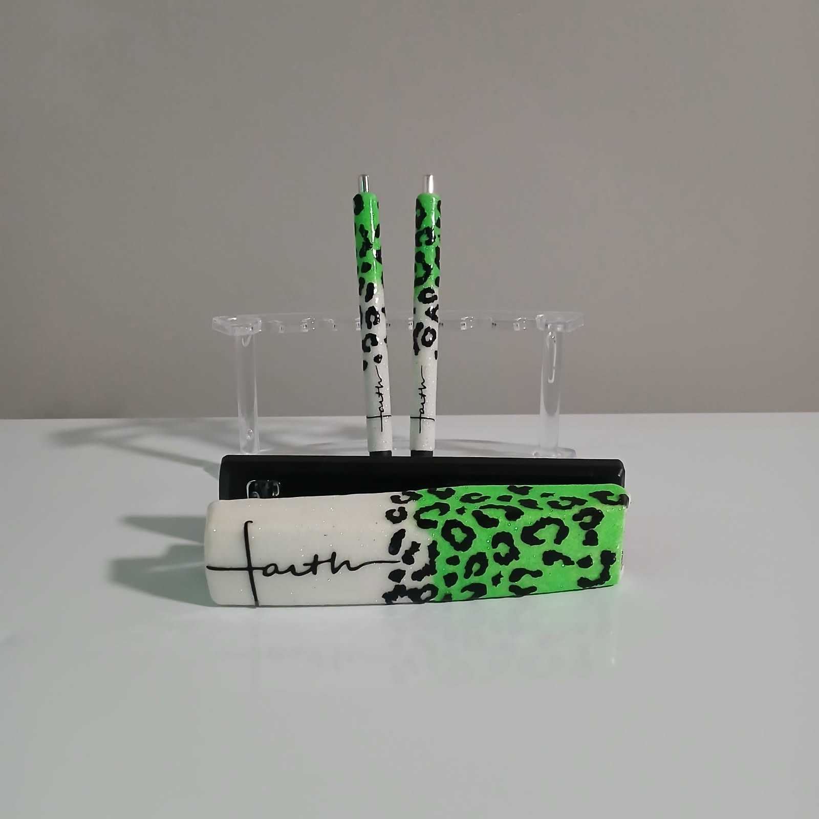 Two decorated pens stand upright on a clear stand, with a rectangular object featuring a green and black leopard print pattern and handwritten text in front, against a plain background.