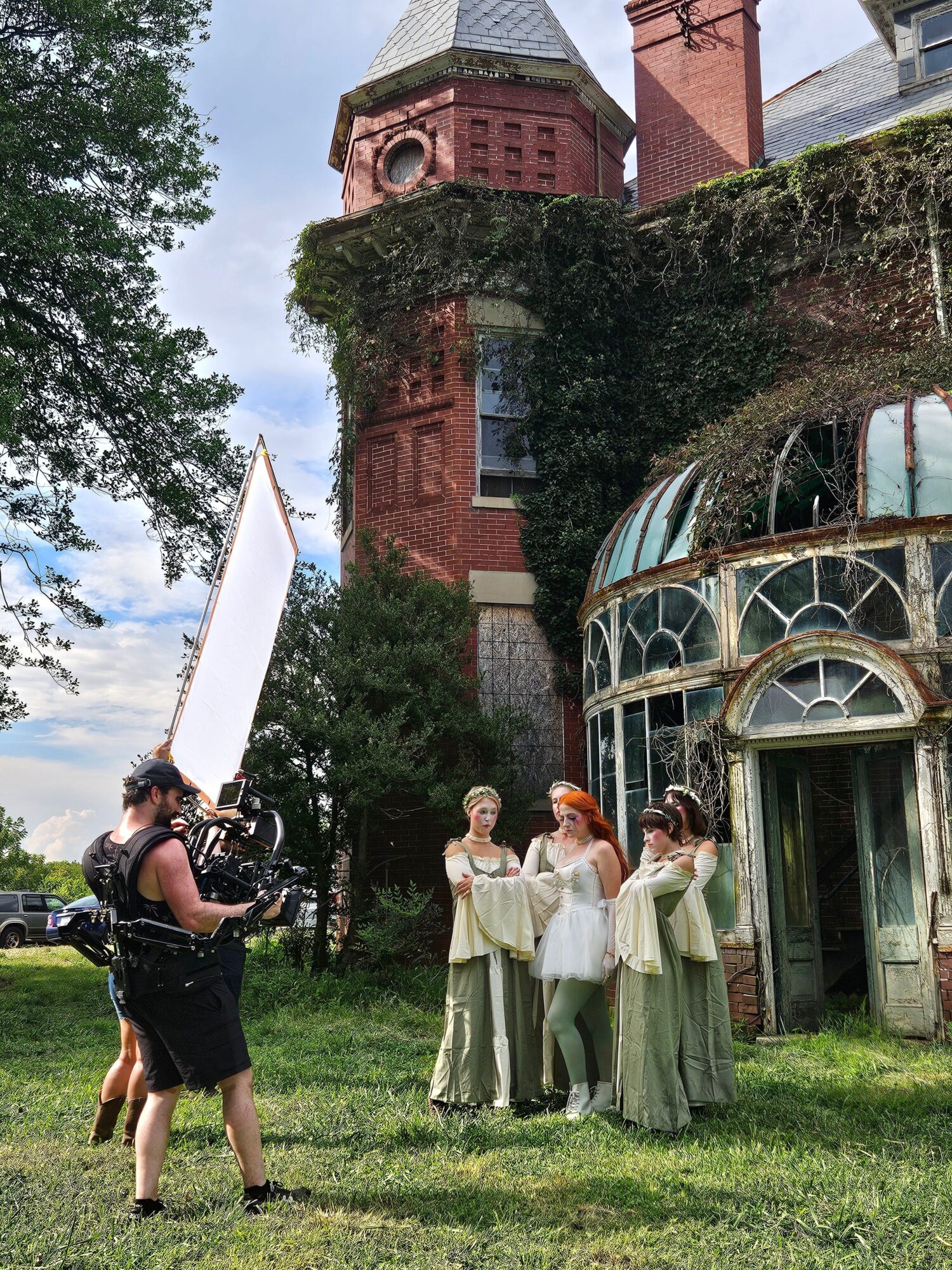 Group of people in vintage clothing standing on grass near a glass greenhouse and large tree, with a person holding a camera.