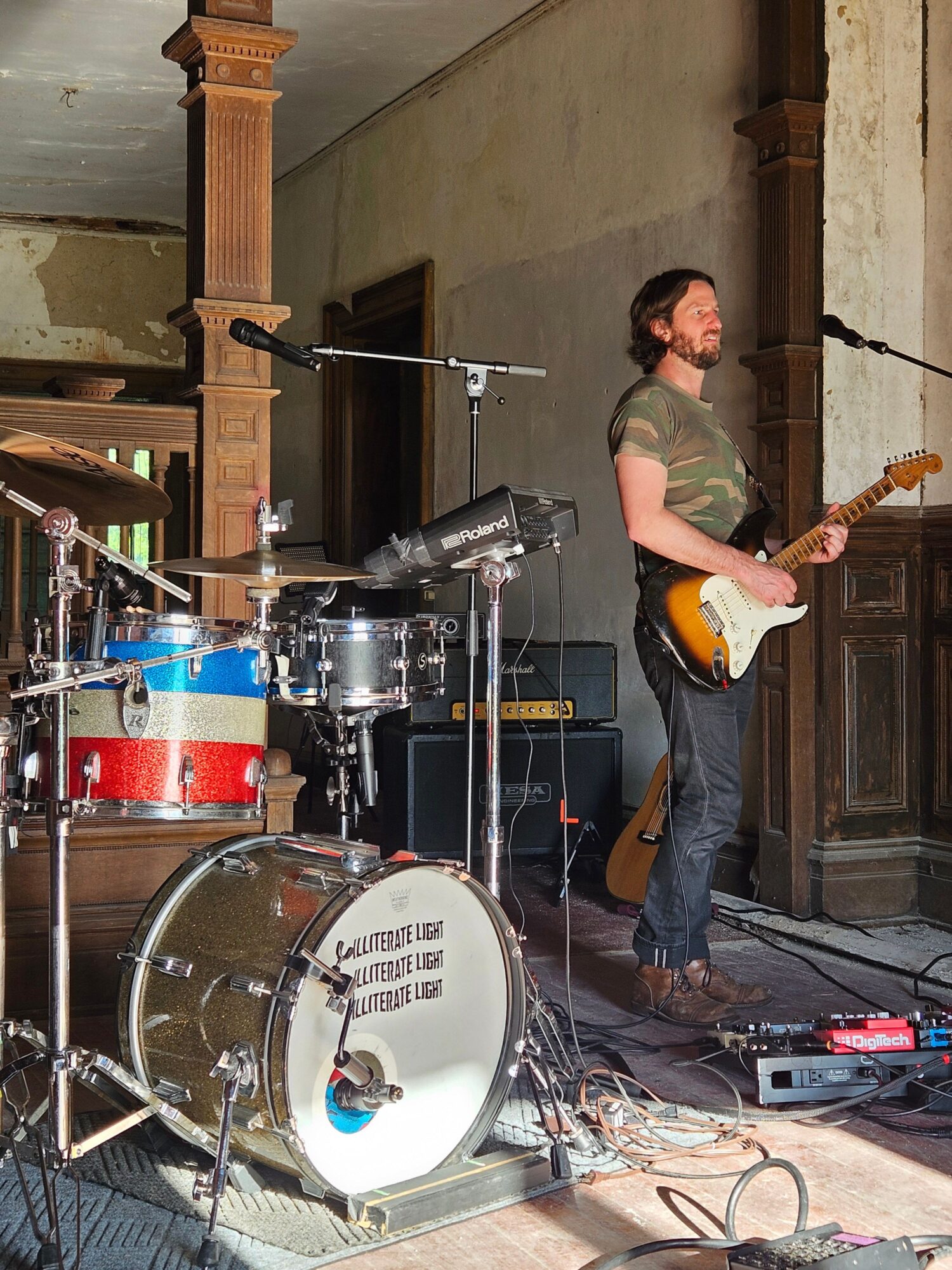Musician playing electric guitar on stage with drum set and equipment, indoor setting, wooden paneling, and cables visible.