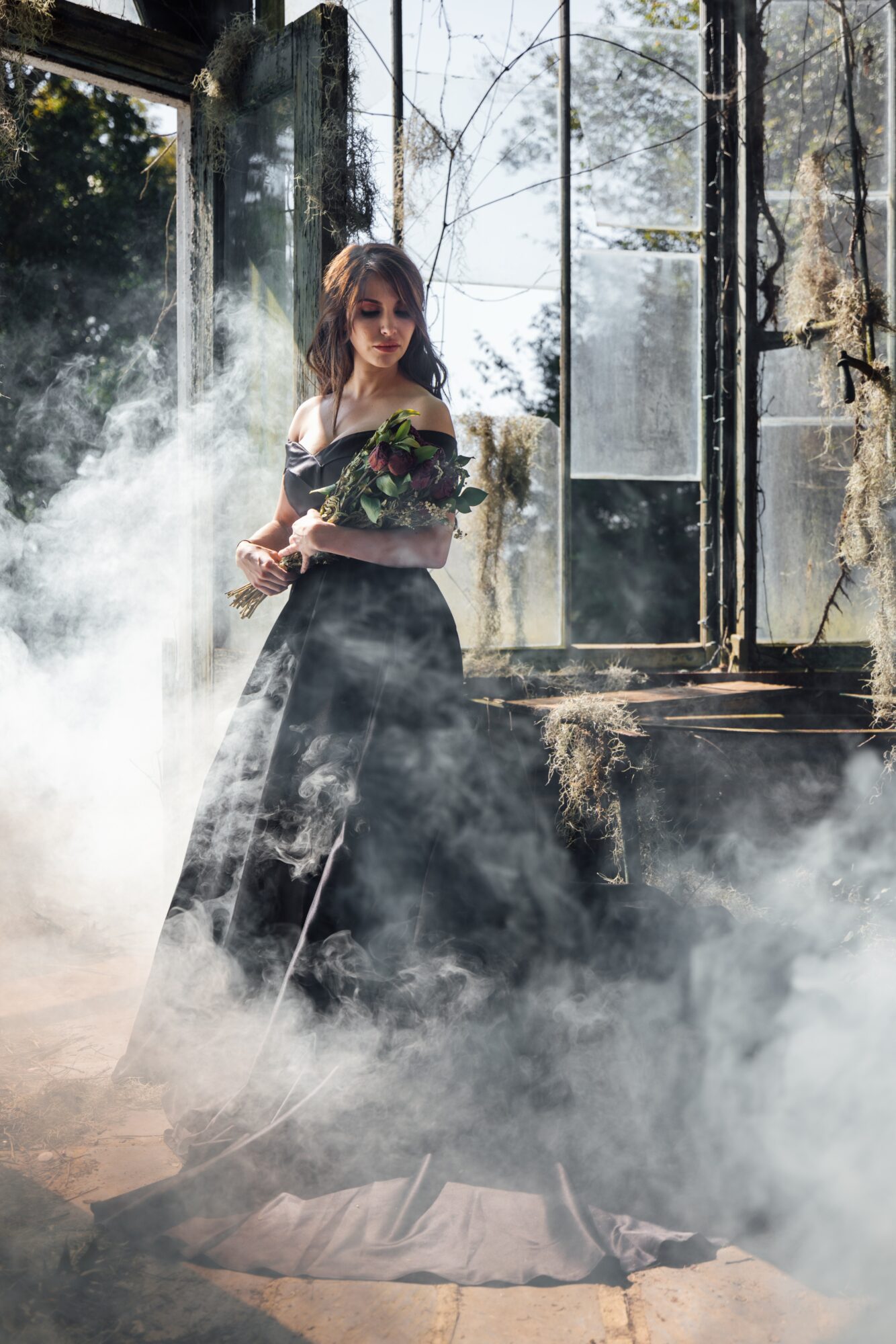 Woman in a black dress holding a bouquet, standing in a misty greenhouse with glass panels and trees outside.