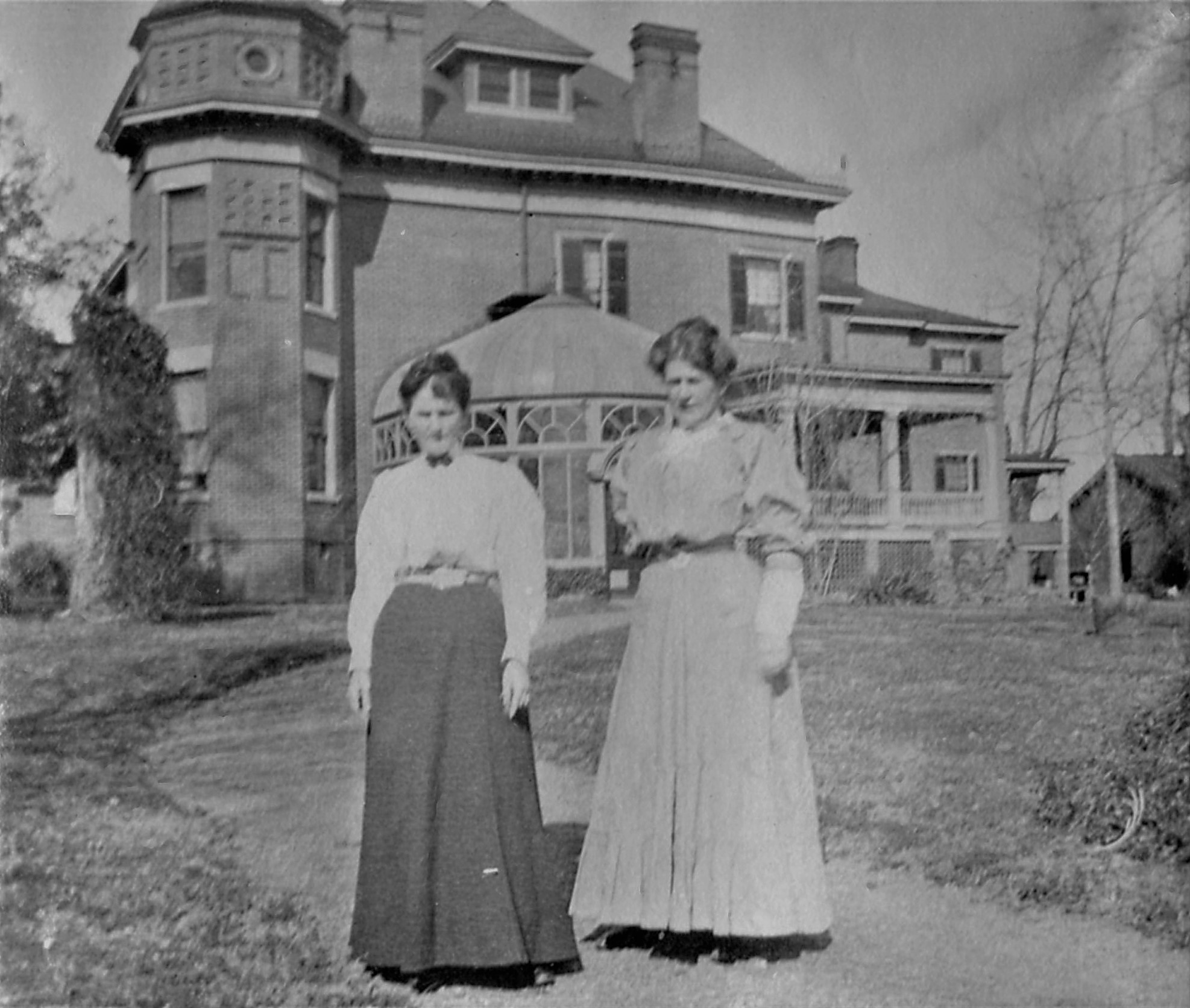 Two women stand outside a large historic house with a turret, trees, and a lawn in the background.