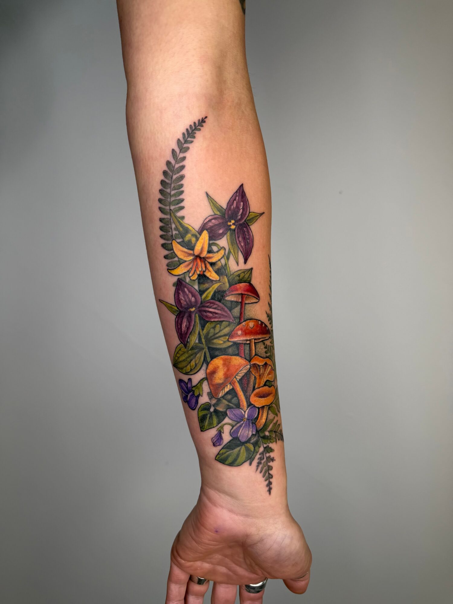 Colorful botanical tattoo with flowers, mushrooms, and leaves on person's forearm, vertical orientation.