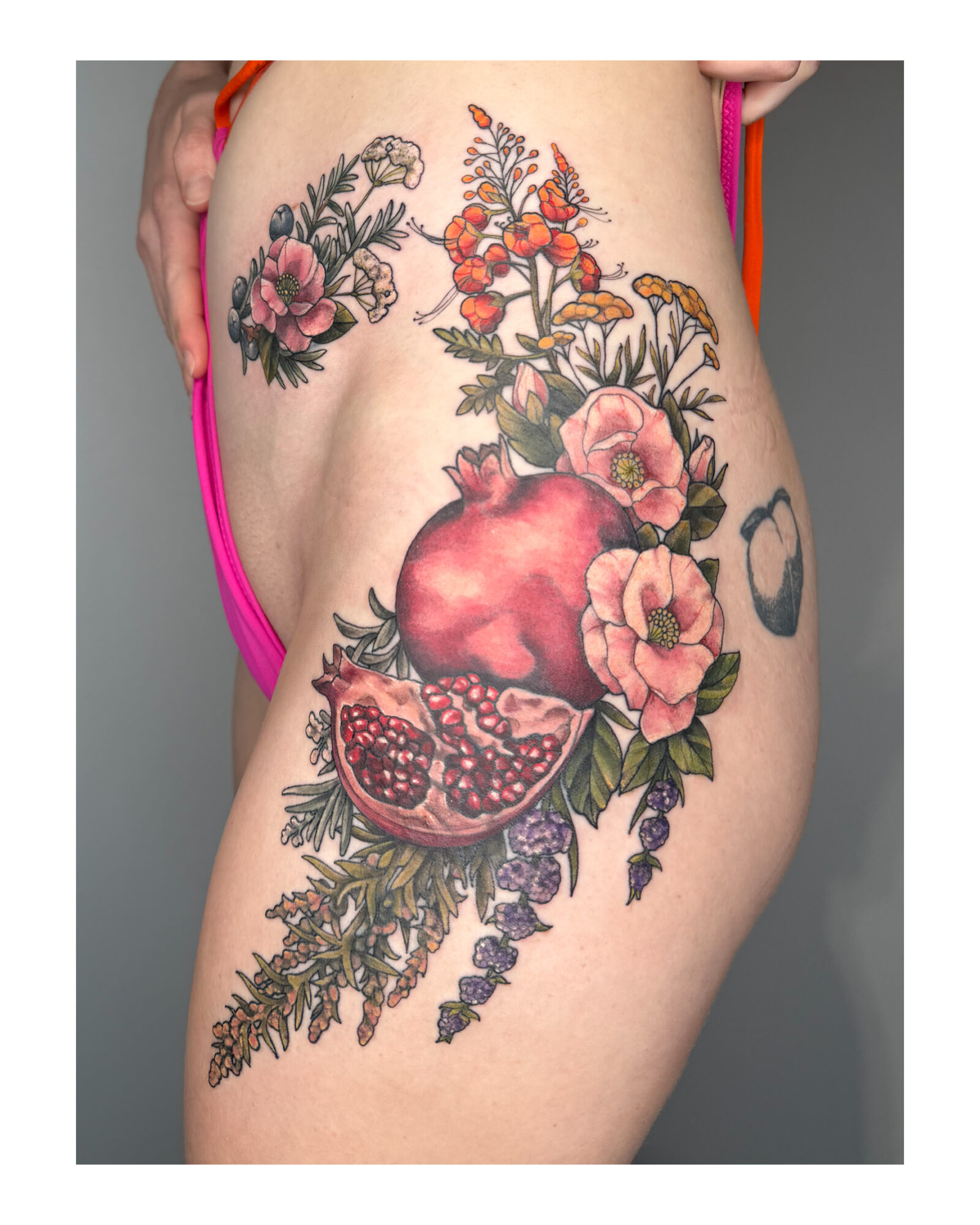 Tattoo of pomegranate, flowers, and leaves on person's thigh, with pink and orange clothing visible.
