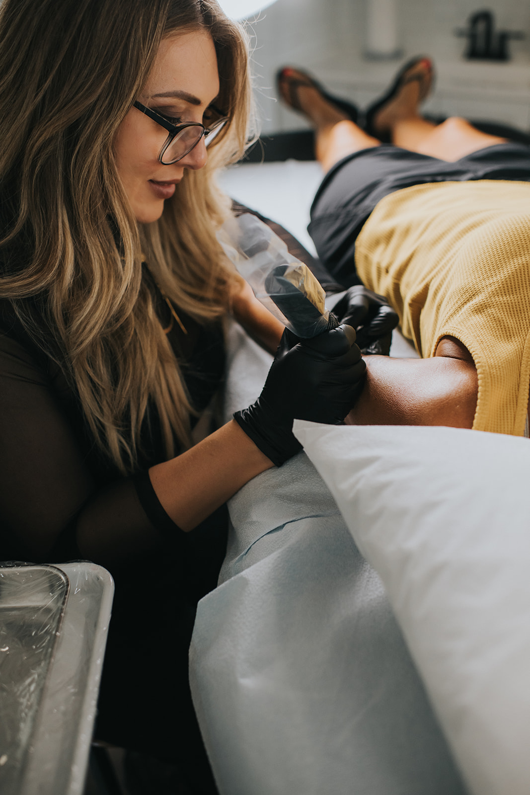 Person with glasses and gloves drawing on a person's arm in a room with a bed and white walls.