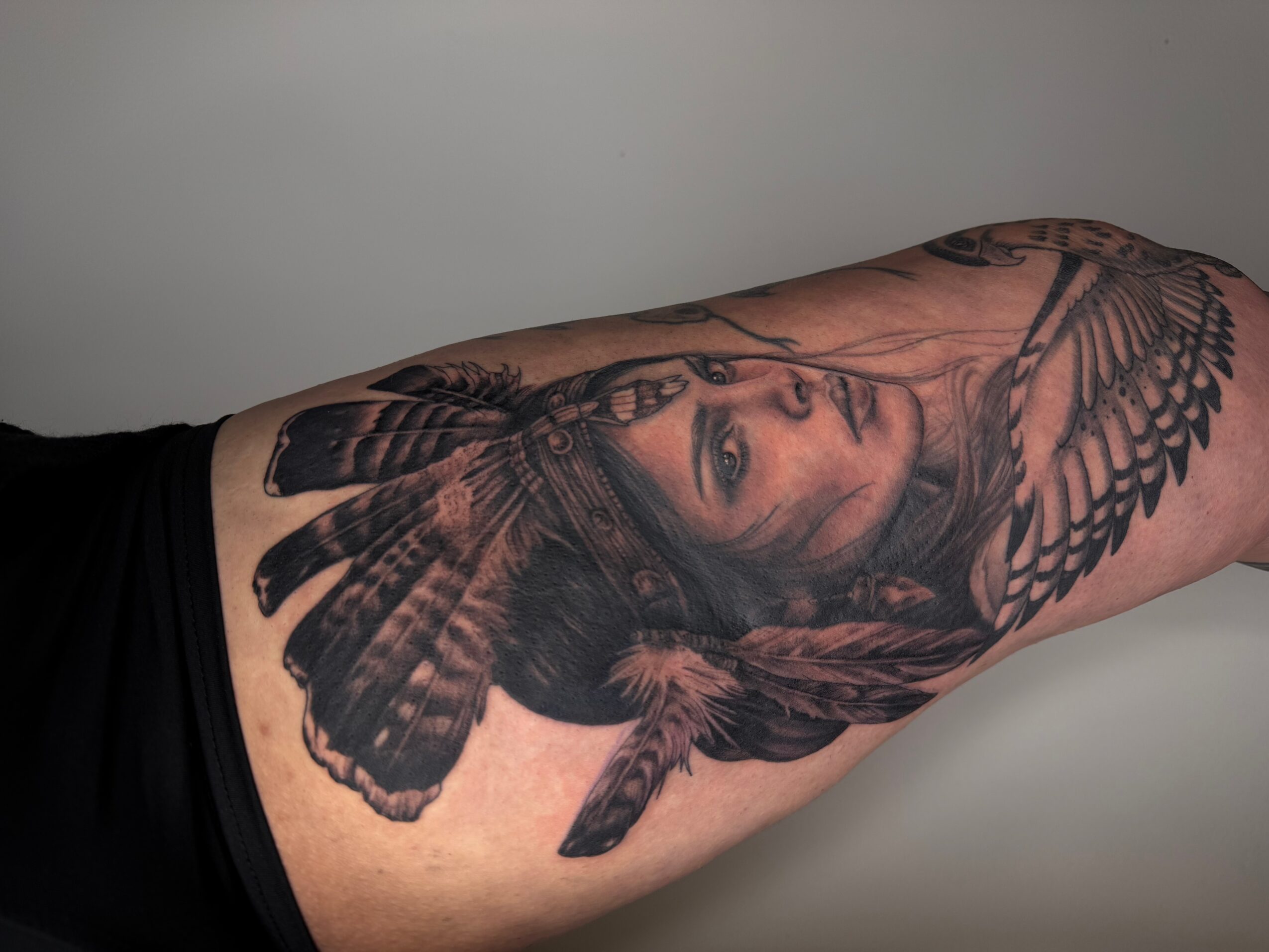 Tattoo of a woman's face with wings and feathers on arm, detailed shading and realistic style.
