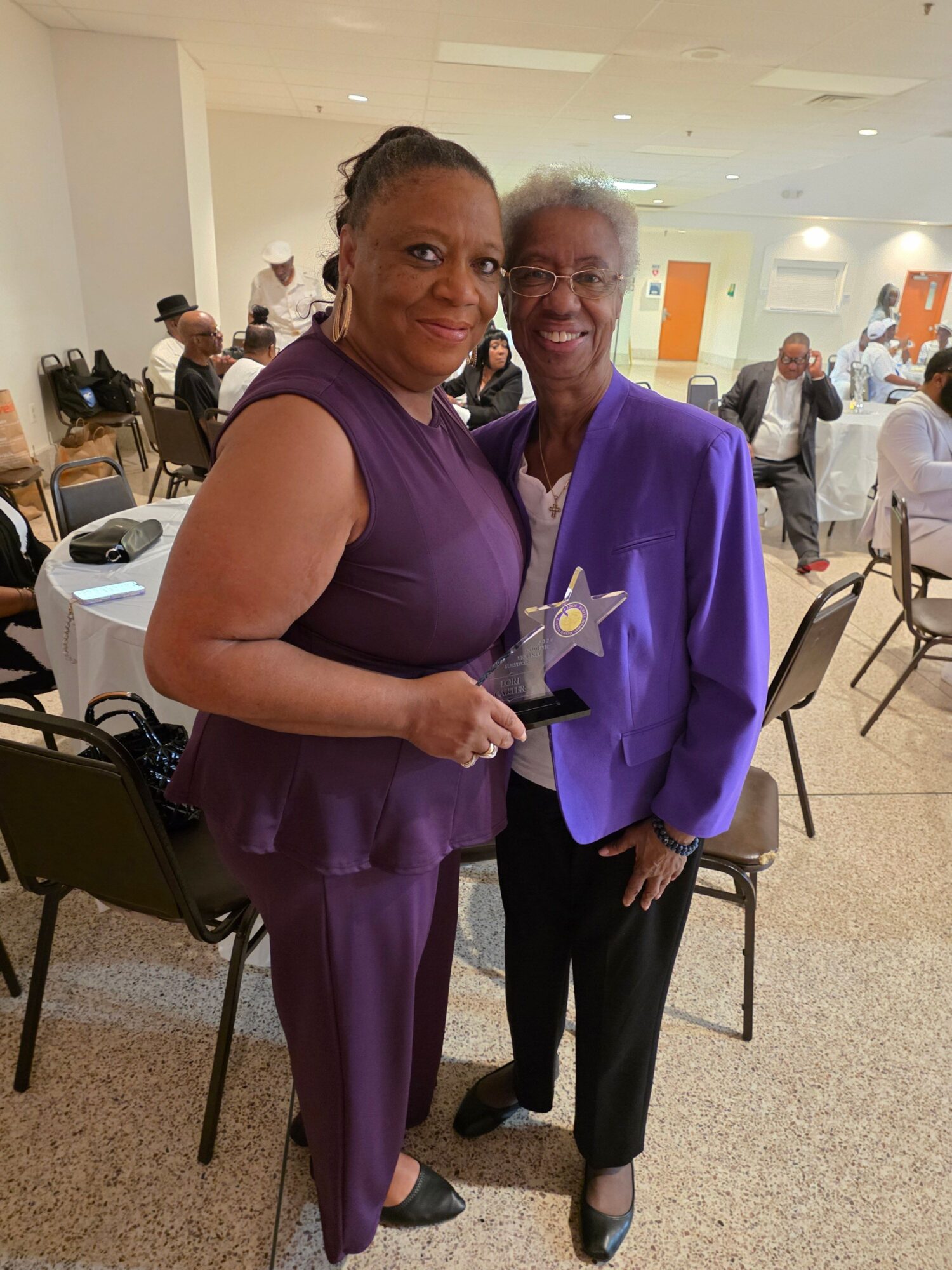 Two women standing indoors, one holding a star-shaped award, with chairs and people in the background.