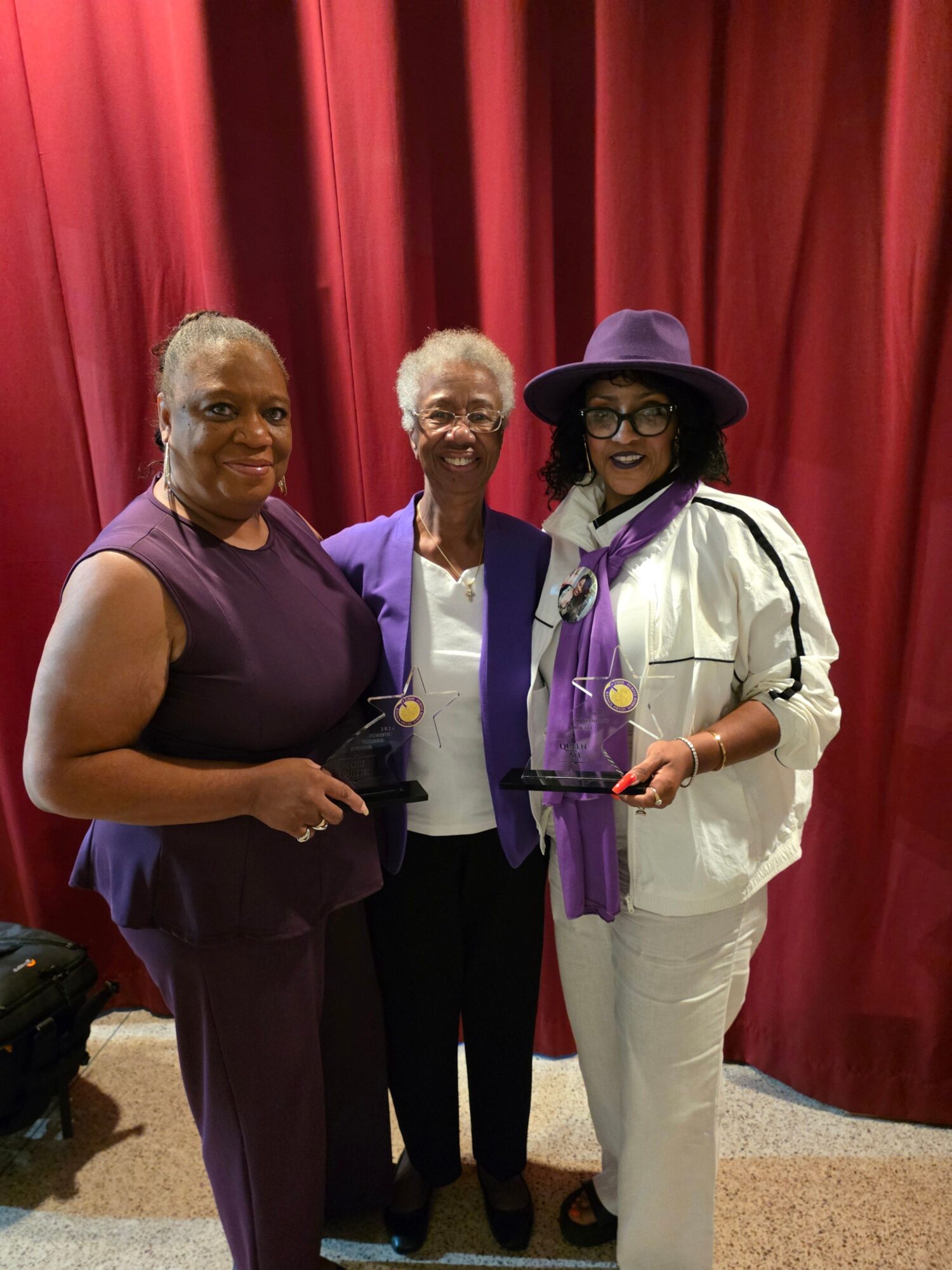 Three women standing together in front of a red curtain, two holding microphones, smiling.