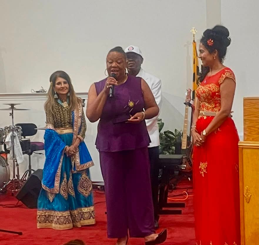 Four women standing on a stage, one speaking into a microphone, dressed in colorful traditional attire, with a white wall background.