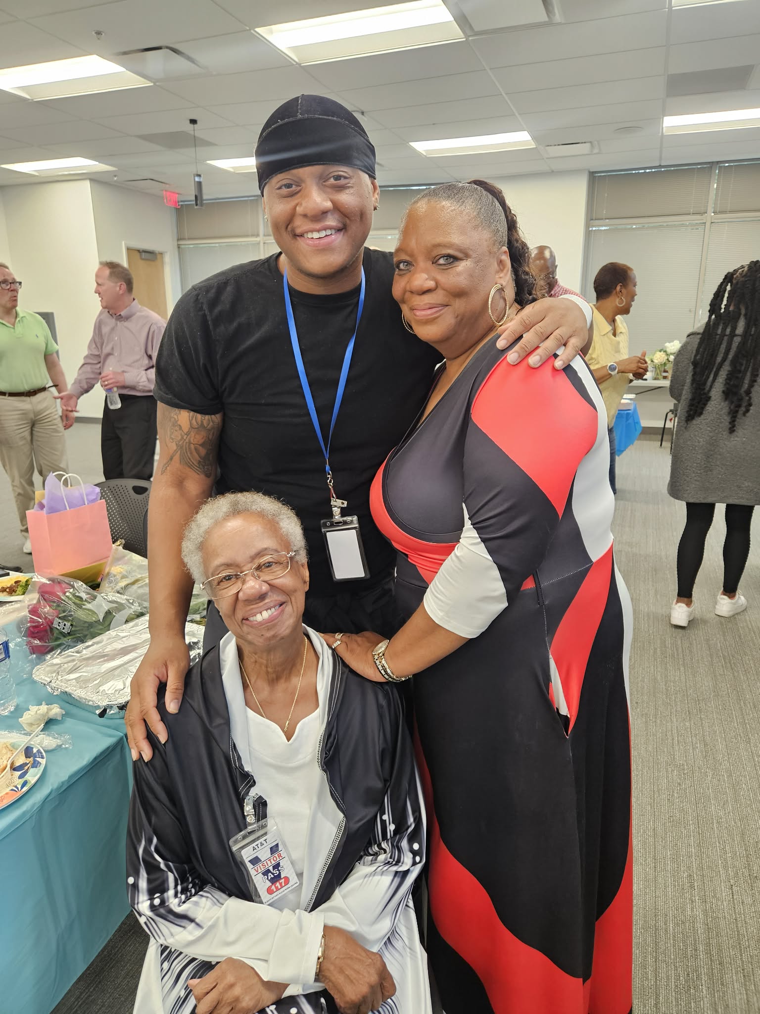 Three people smiling and posing together indoors, with a table of flowers and other people in the background.