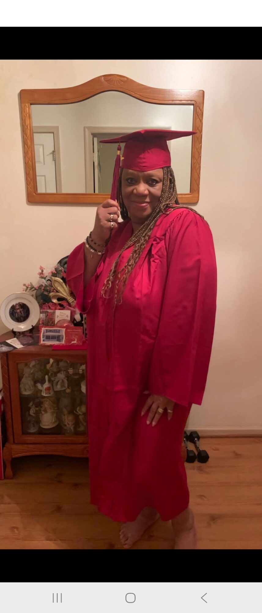 Person in a red graduation gown and cap giving a thumbs-up, standing in front of a mirror and furniture.