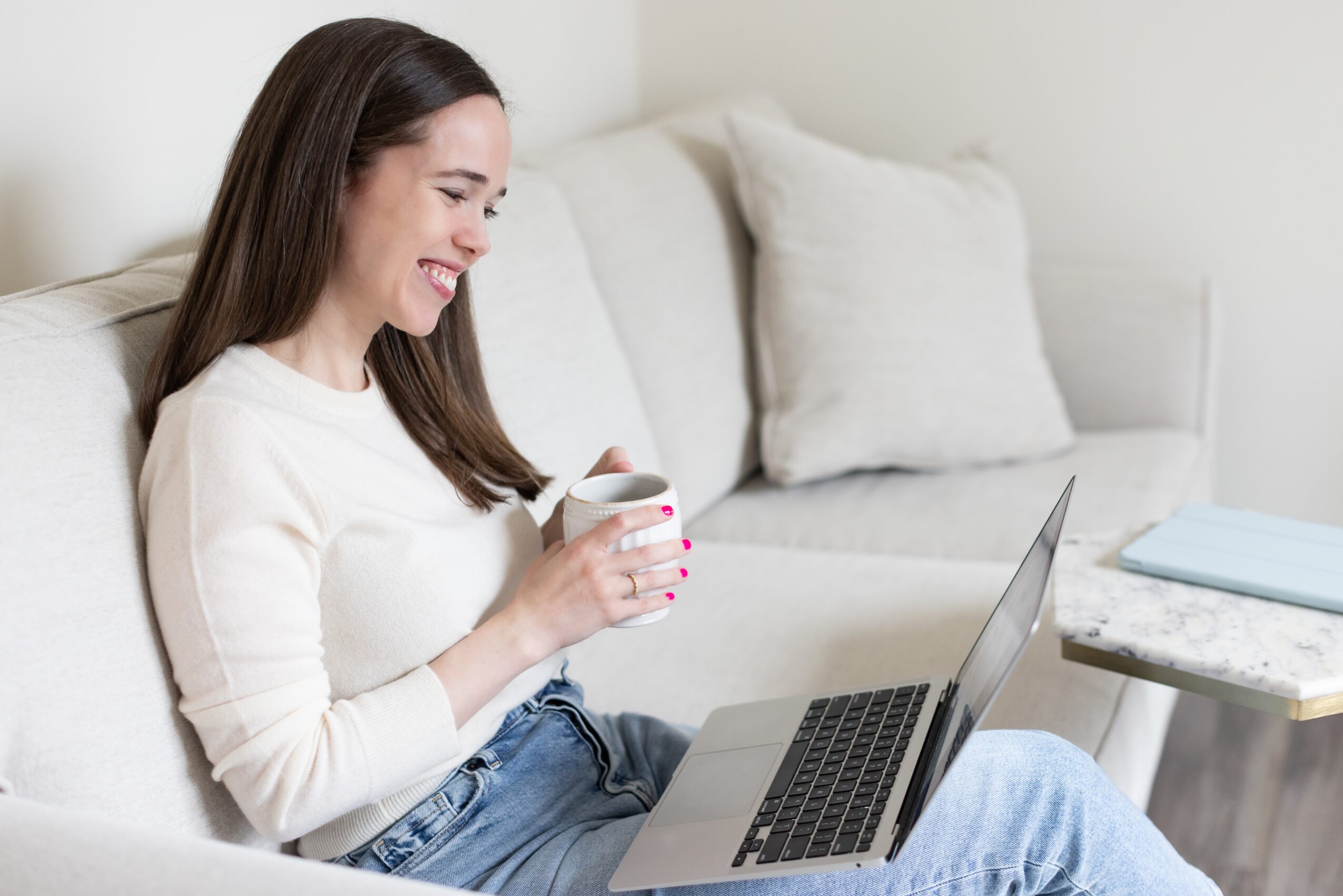 Woman sitting on a sofa, smiling, holding a mug, using a laptop, with pillows and a table nearby