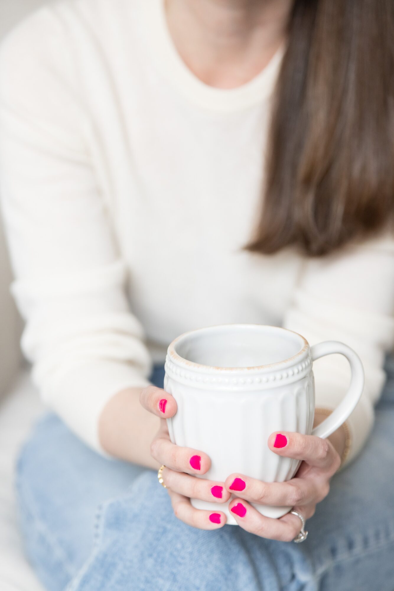 Person holding a white mug with pink nail polish, wearing a white sweater and blue jeans.