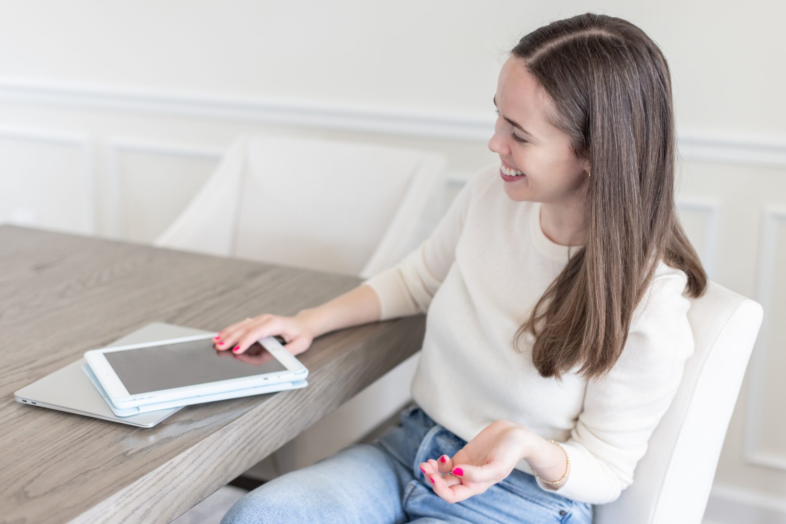 Woman sitting at a table with a laptop and notebook, smiling and holding a pen, in a bright room.