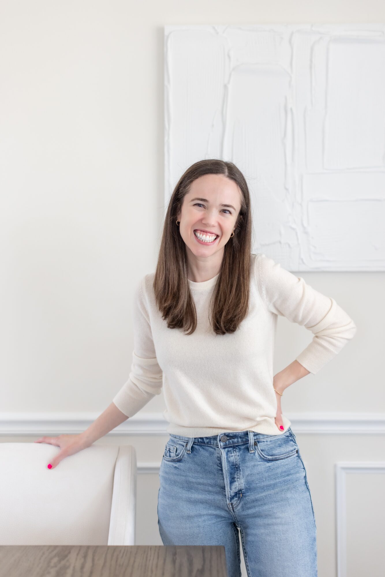 Smiling woman with long brown hair in a white sweater and jeans, standing indoors near a white wall and artwork.