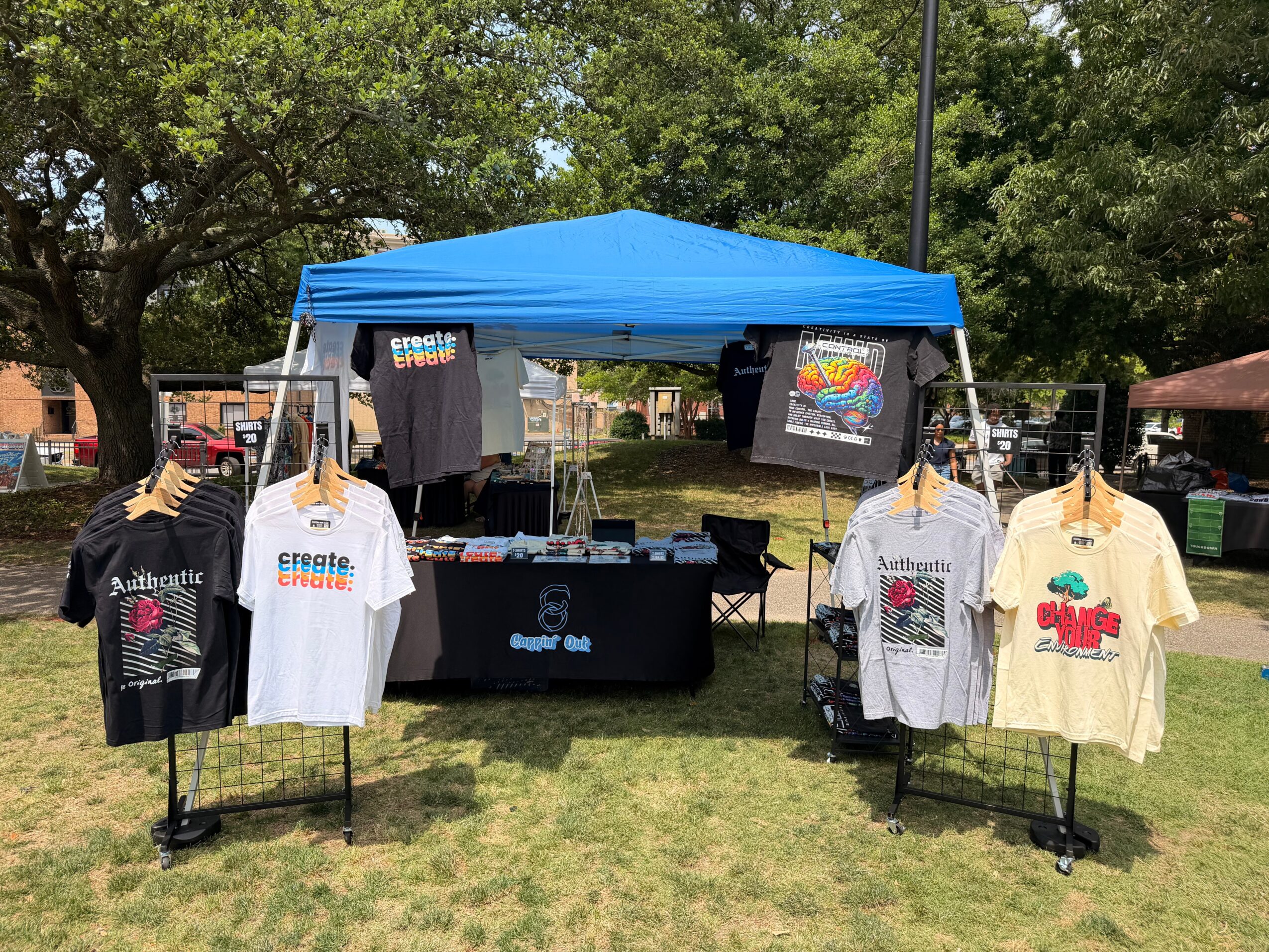 Outdoor booth with T-shirts on display under a blue canopy, set on grass with trees in the background.
