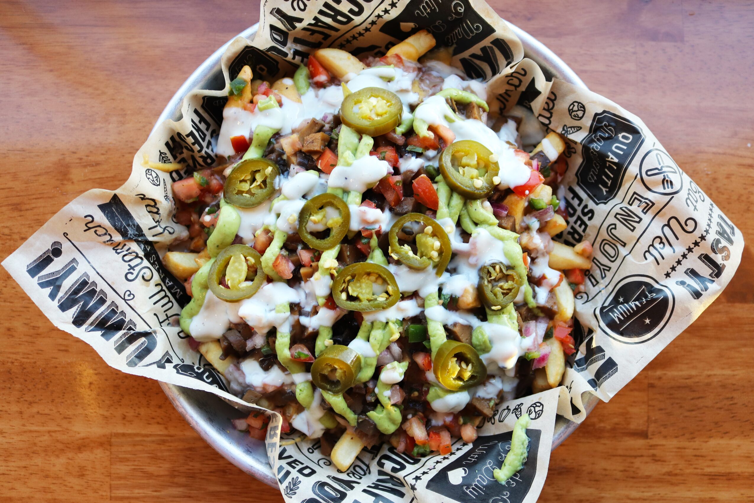 Bowl of nachos topped with melted cheese, jalapeños, and diced vegetables on a wooden table.