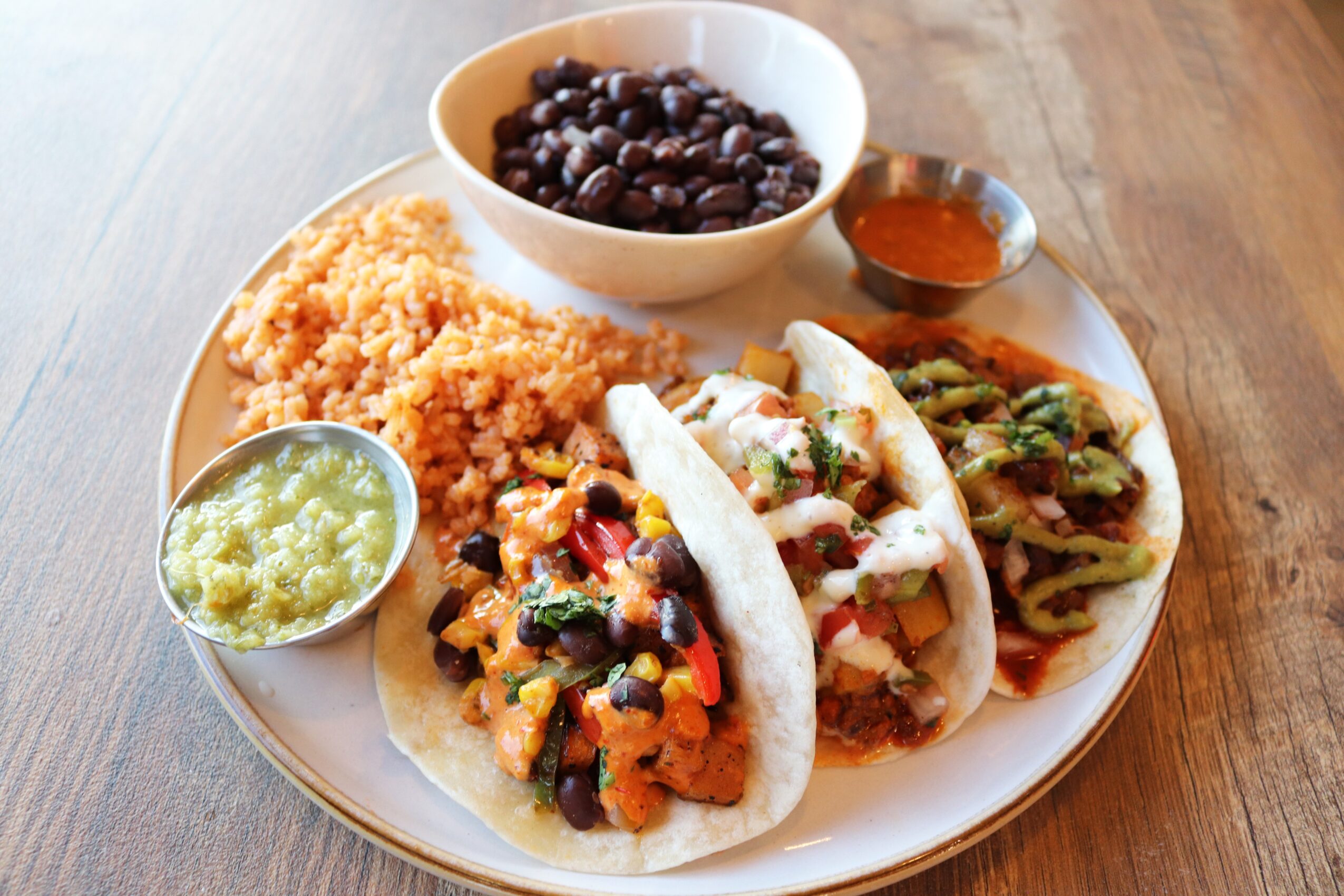 Plate with rice, beans, guacamole, two tacos with toppings, and side dishes on a wooden table.