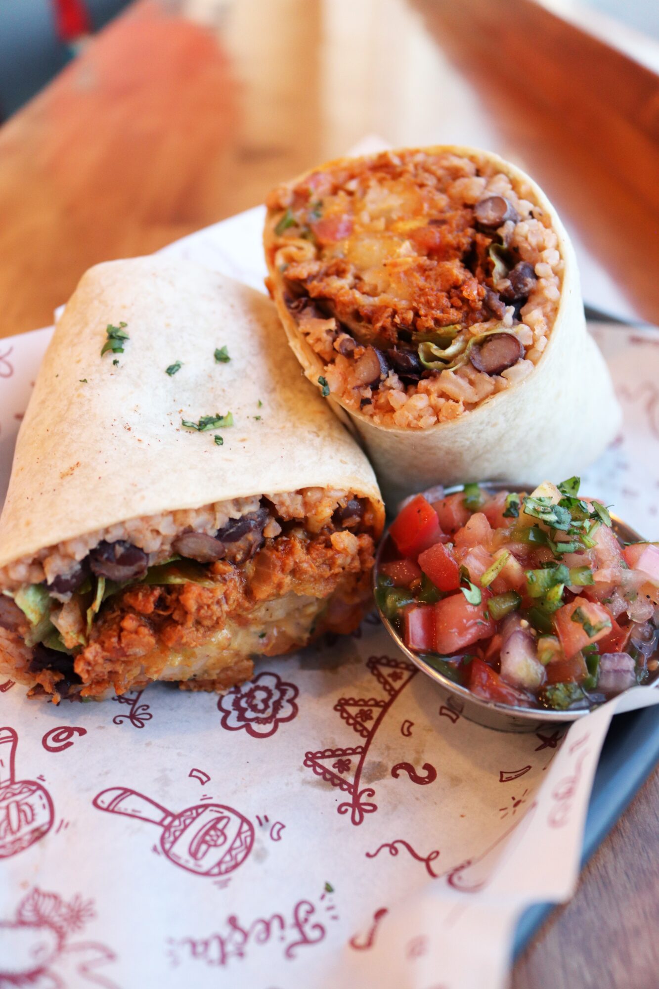 Close-up of a burrito cut in half, showing rice, beans, and vegetables, with salsa on the side, on paper with drawings.