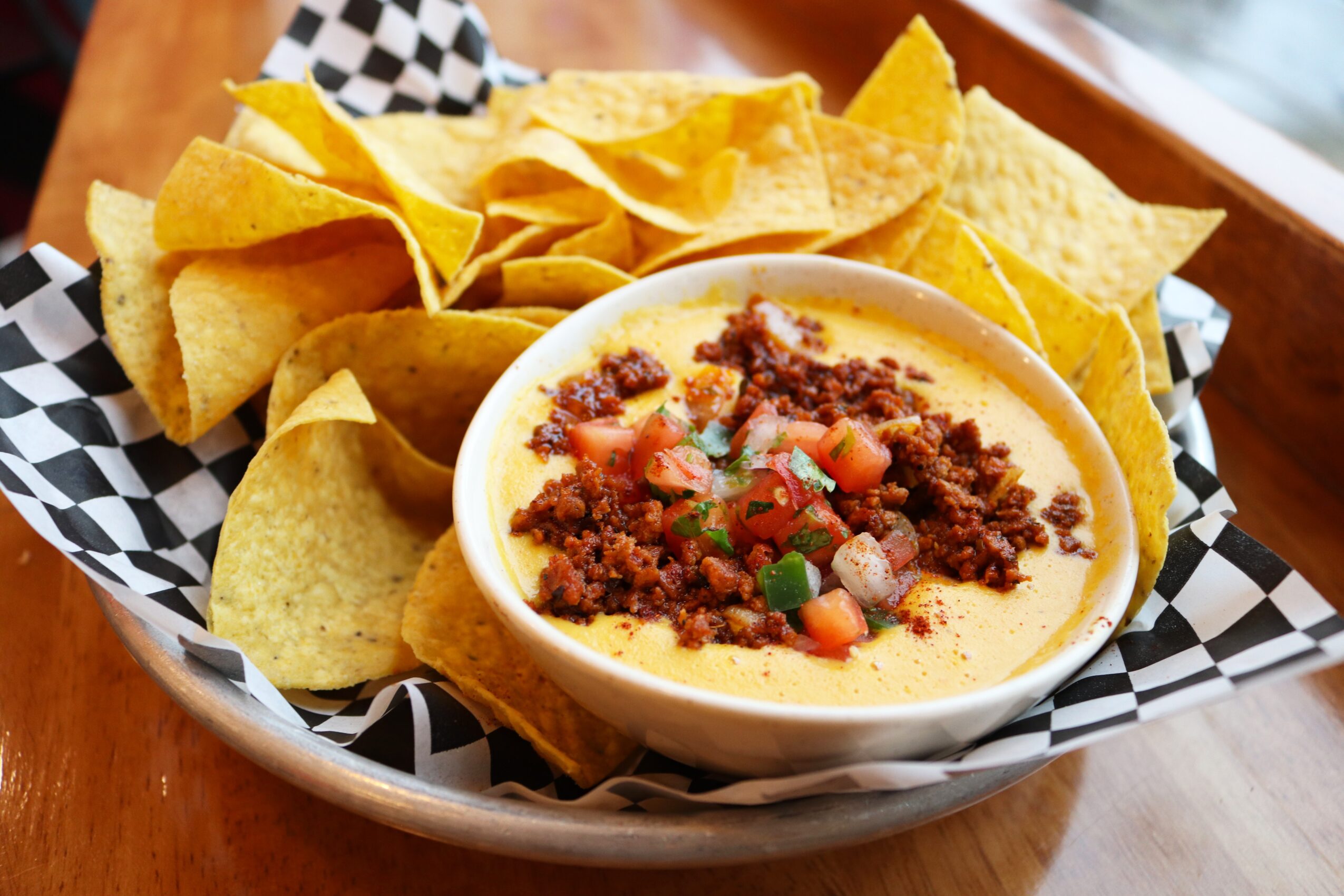Bowl of nacho cheese with ground beef, tomatoes, and green onions, surrounded by tortilla chips in a basket.