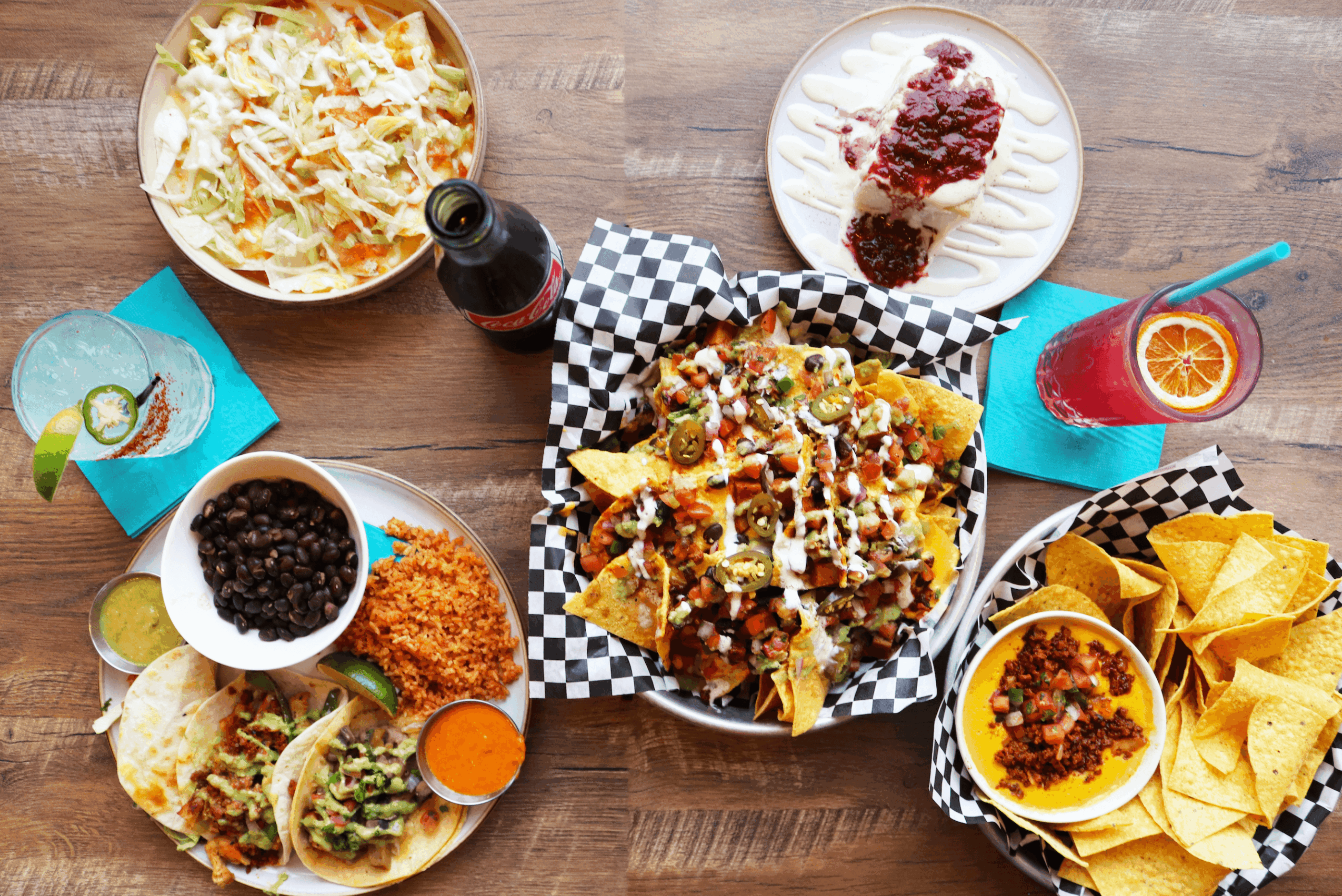 Top-down view of a table with nachos, chips with salsa, a salad, a drink, a dessert, and a bowl of black beans.