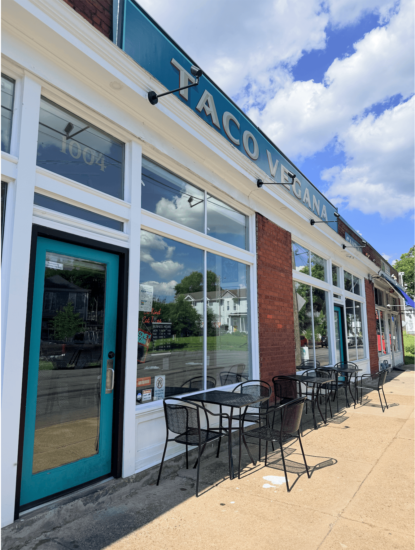Exterior of Taco Y Juana restaurant with outdoor seating, large windows, and a blue sign, under a partly cloudy sky.