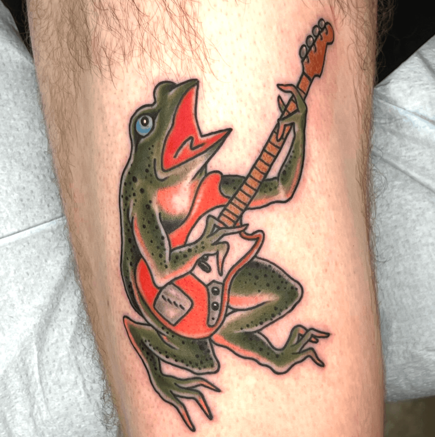 A frog playing an electric guitar, with detailed features and vibrant colors, on a person's leg.