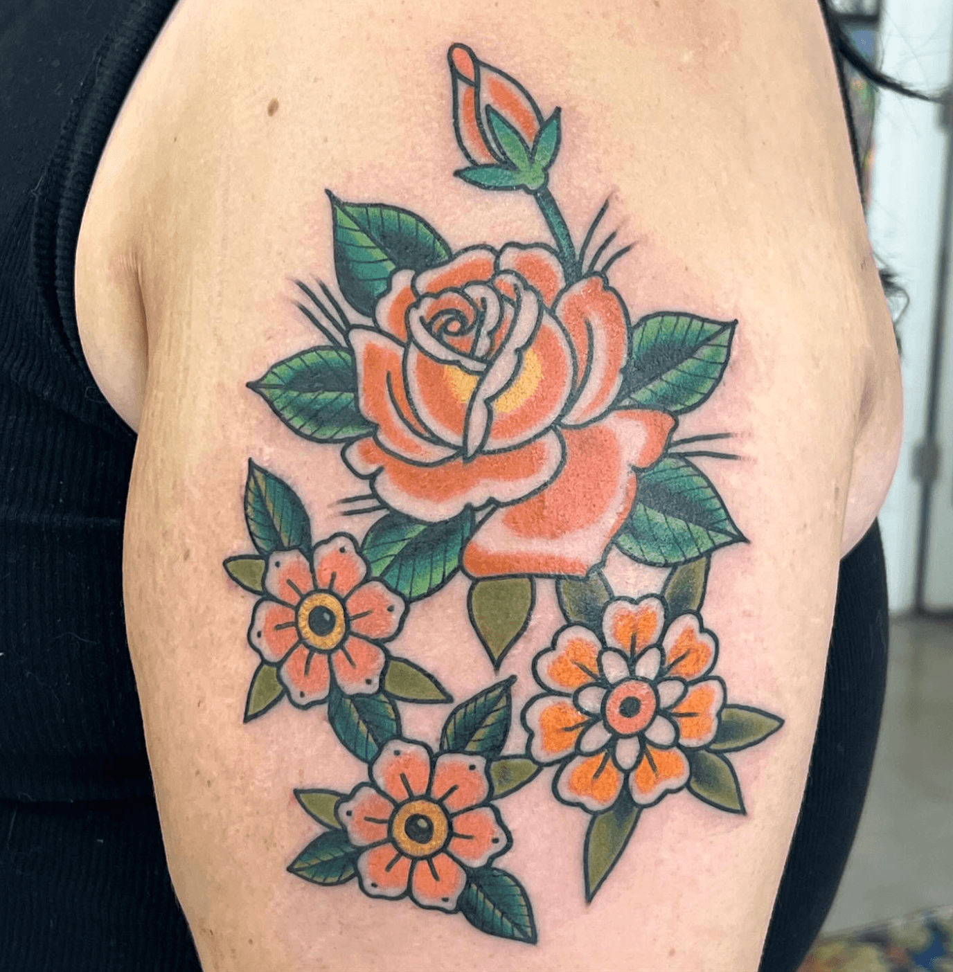 Colorful rose and flower tattoo on upper arm with green leaves, outlined in black, in traditional style.