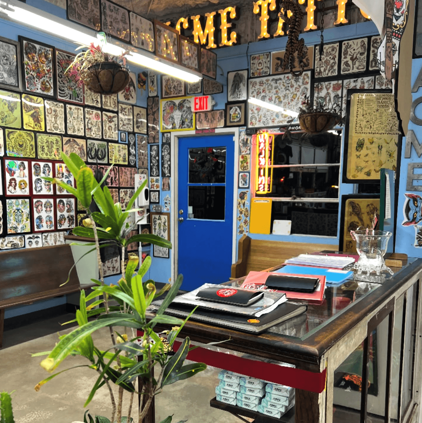 Interior of a tattoo shop with framed artwork on walls, a blue door, and a counter with notebooks and a glass jar.