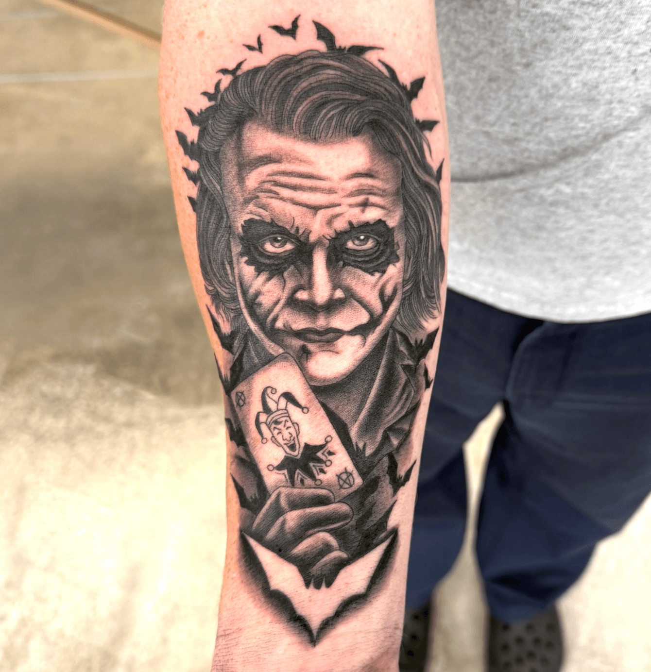 Tattoo of a man with a sinister expression, holding a card, with bats flying above, in black and gray ink.