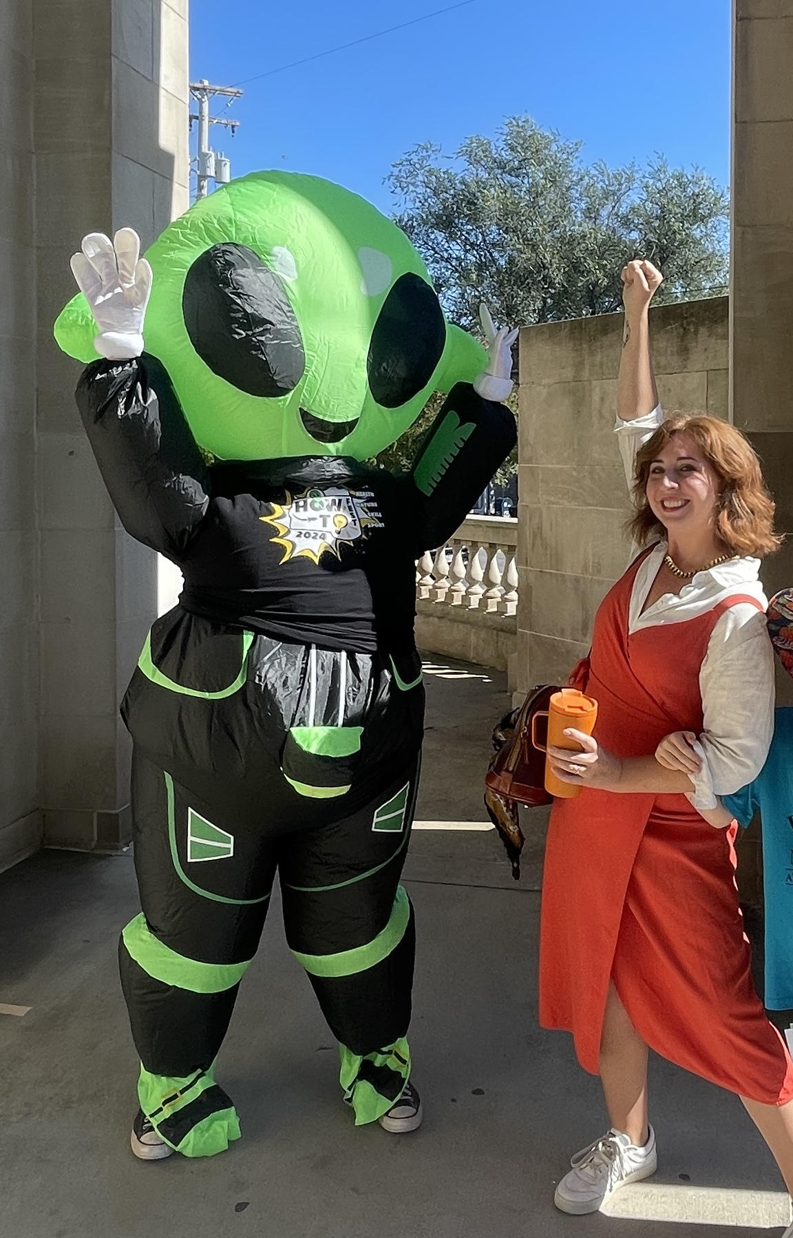 Person in an alien costume waving next to a woman holding a drink outdoors, with trees and a building in the background.