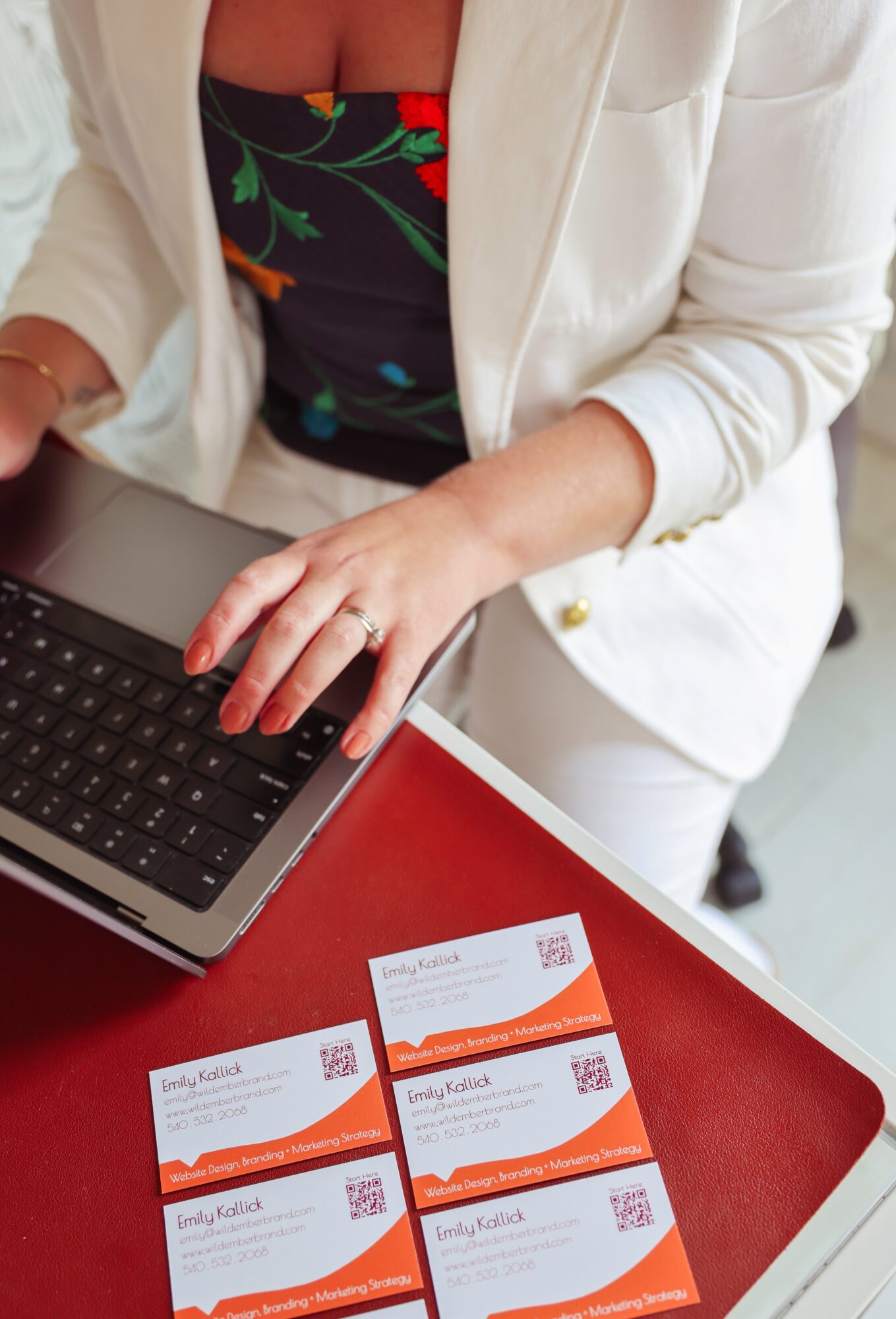 Person in white blazer and floral top working on laptop at red table with four business cards.