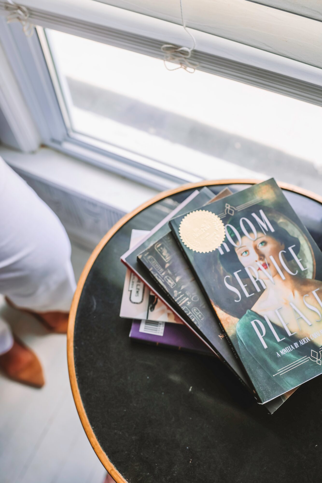 Stack of magazines on a round black table near a window, with a person partially visible nearby.