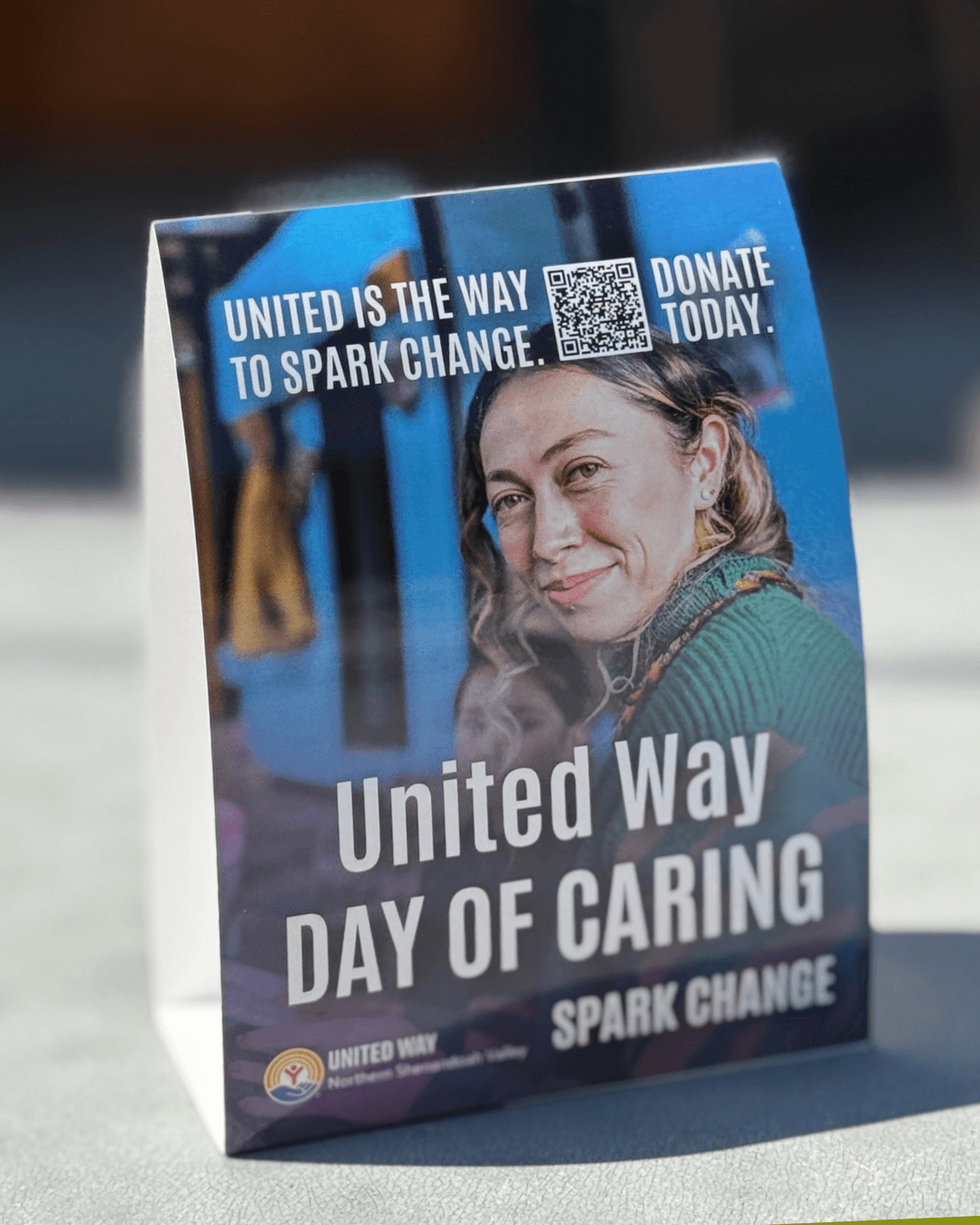 A sign with a woman smiling, promoting United Way Day of Caring and Spark Change, with a QR code and donation message.