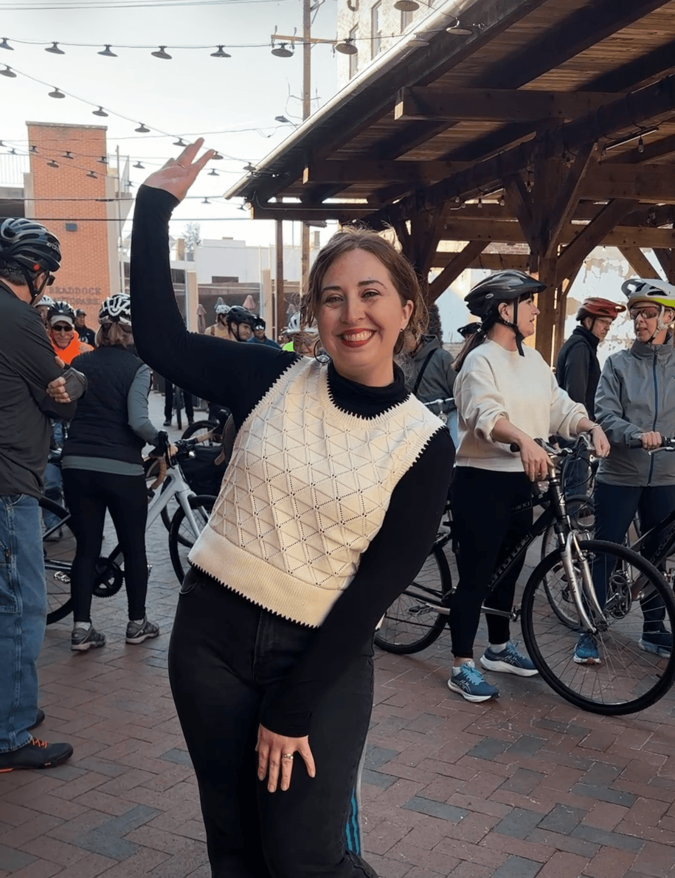 Young woman smiling and waving at outdoor gathering with people and bicycles in background.