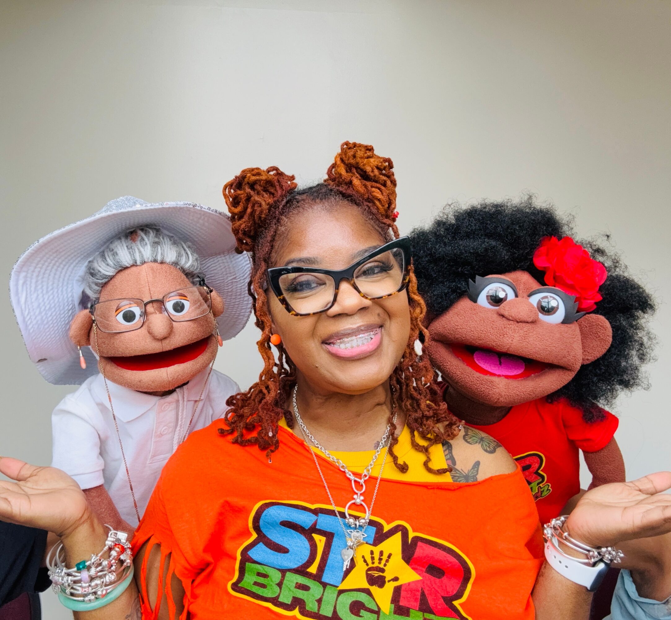 Woman with glasses and braided hair smiling, flanked by two puppets with exaggerated features and colorful clothing.