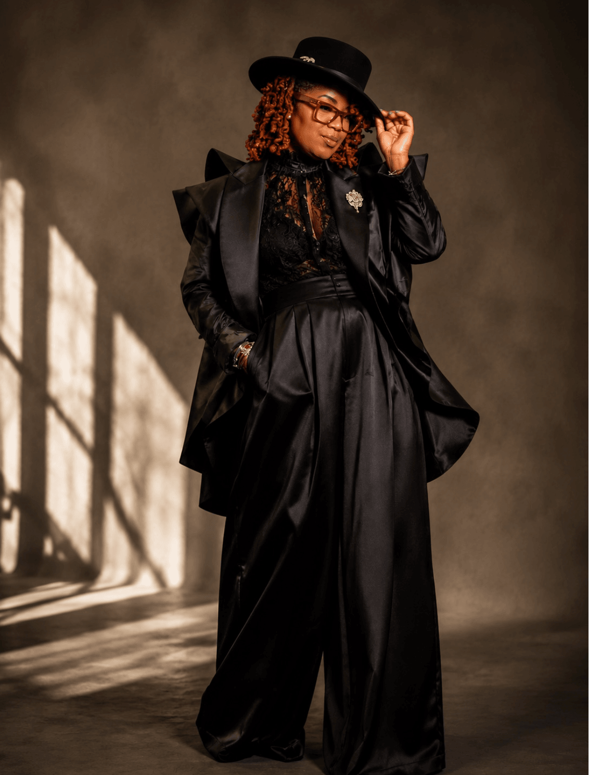 Woman in black outfit, wide-leg pants, jacket, hat, glasses, standing in a dimly lit space with shadows, holding her hat.