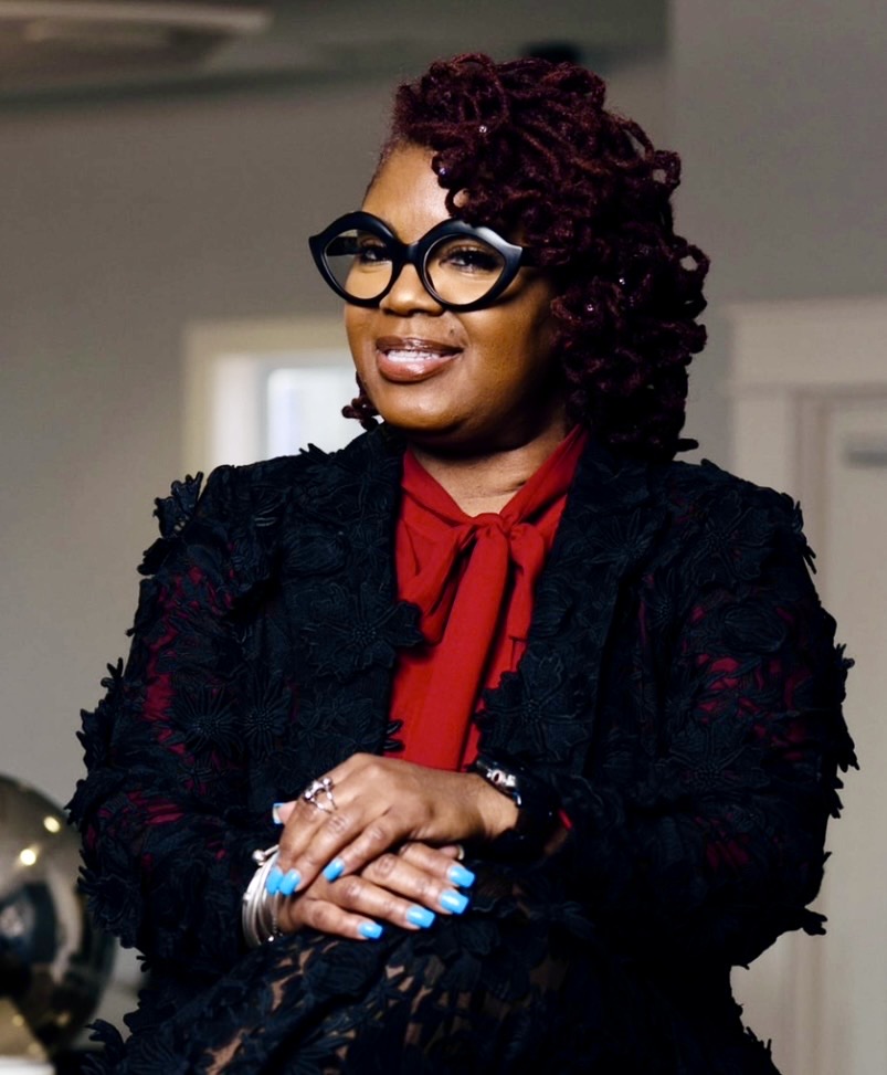 Woman with curly hair, glasses, red blouse, and black jacket, smiling with crossed arms indoors.