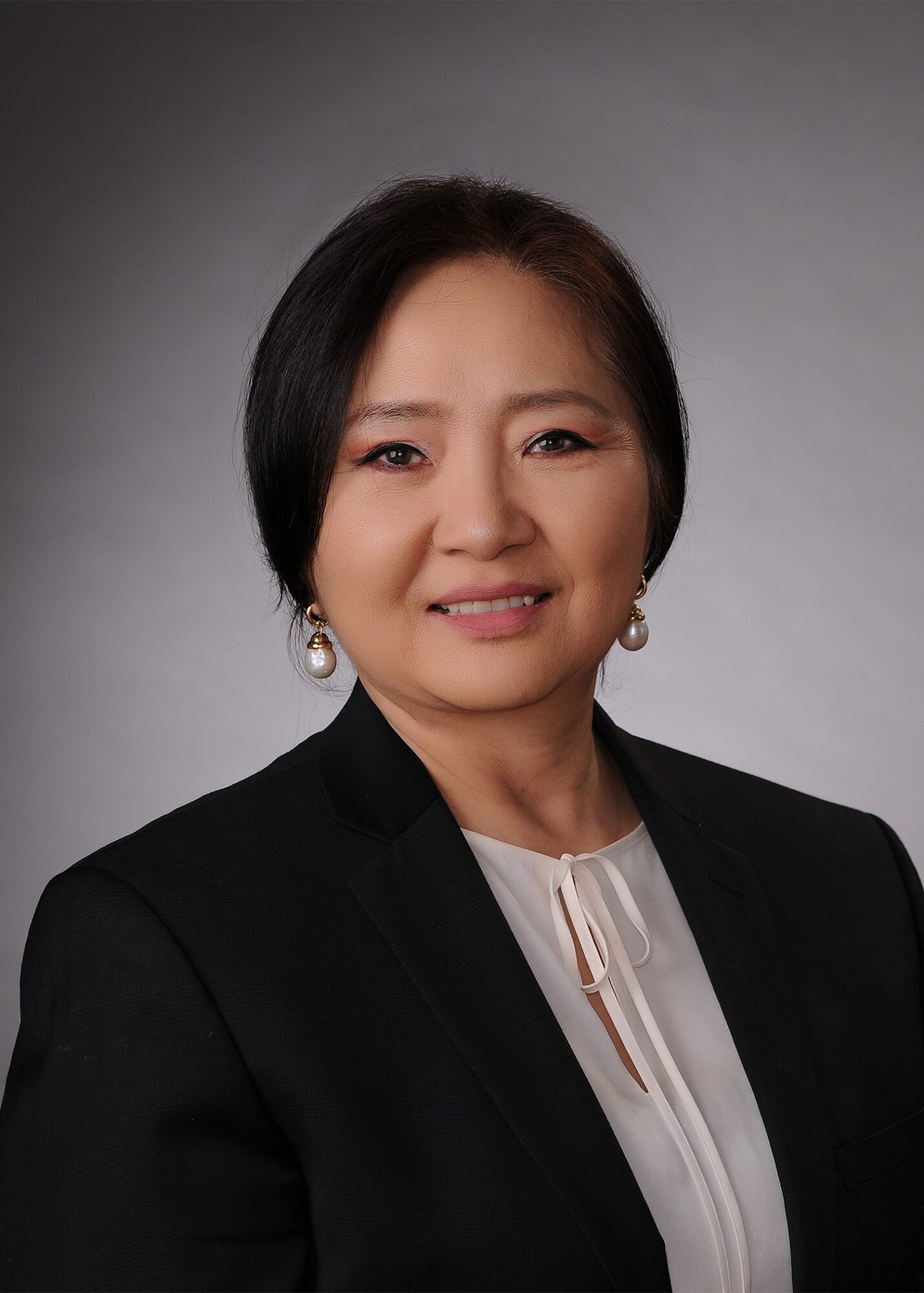 Portrait of a woman with short black hair, wearing a black blazer and white blouse, smiling against a gray background.