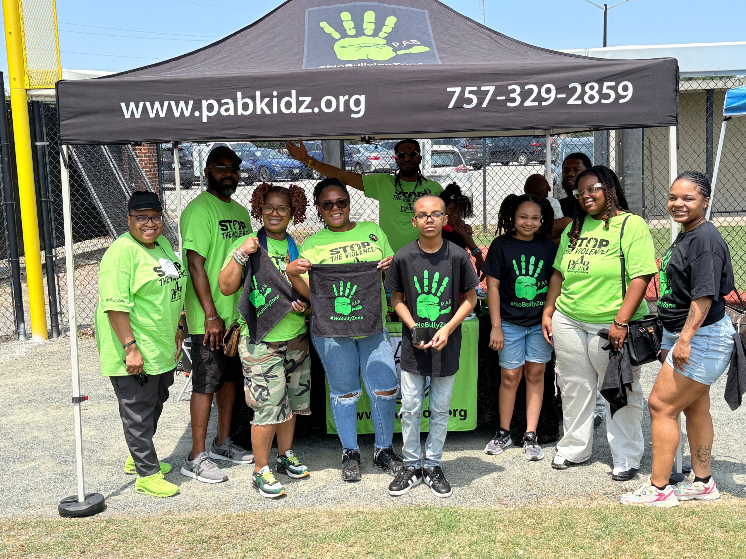 Group of people standing under a black canopy with green hand logo, website, and phone number, outdoors on grass.