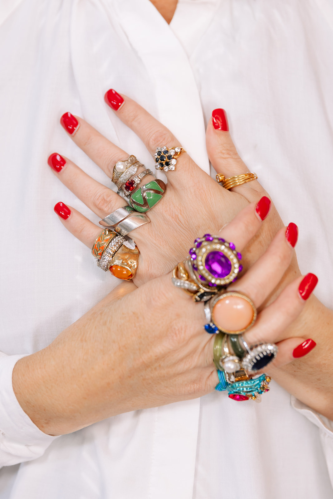 Person's hands with multiple rings and red nail polish, wearing a white garment.