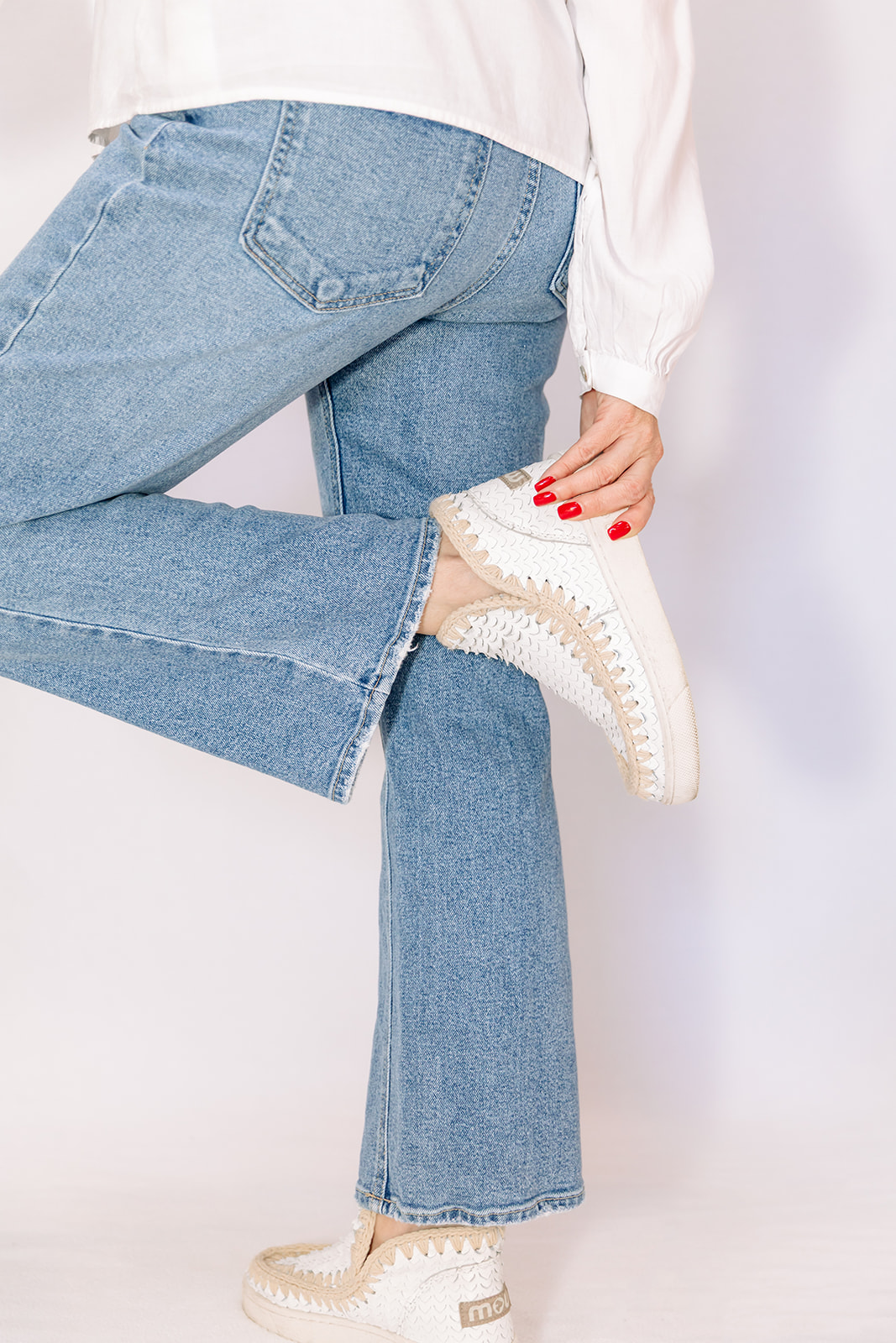 Person wearing light blue jeans and white sneakers, holding their ankle with one hand, against a plain background.