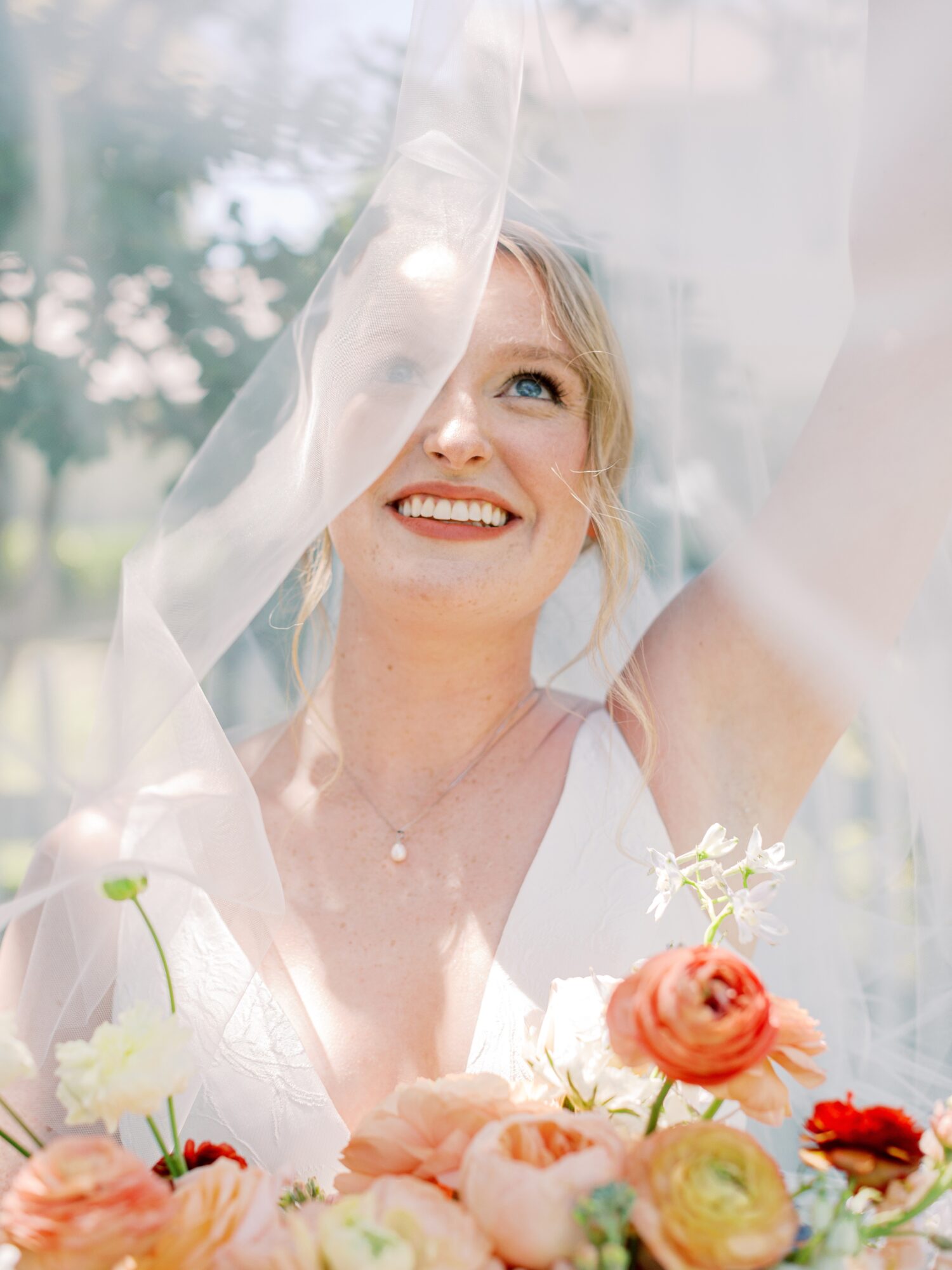 Smiling woman with blonde hair behind sheer fabric, surrounded by flowers, outdoors with trees in background.
