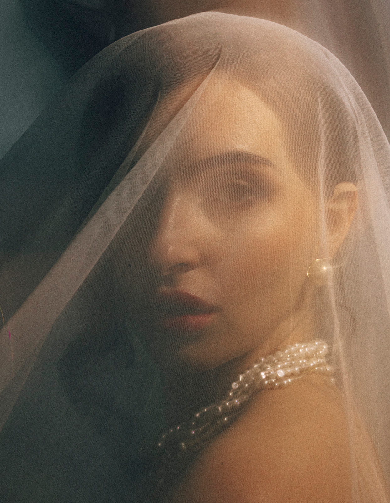 Portrait of a woman with a veil, pearl necklace, and earrings, looking at the camera with soft lighting.